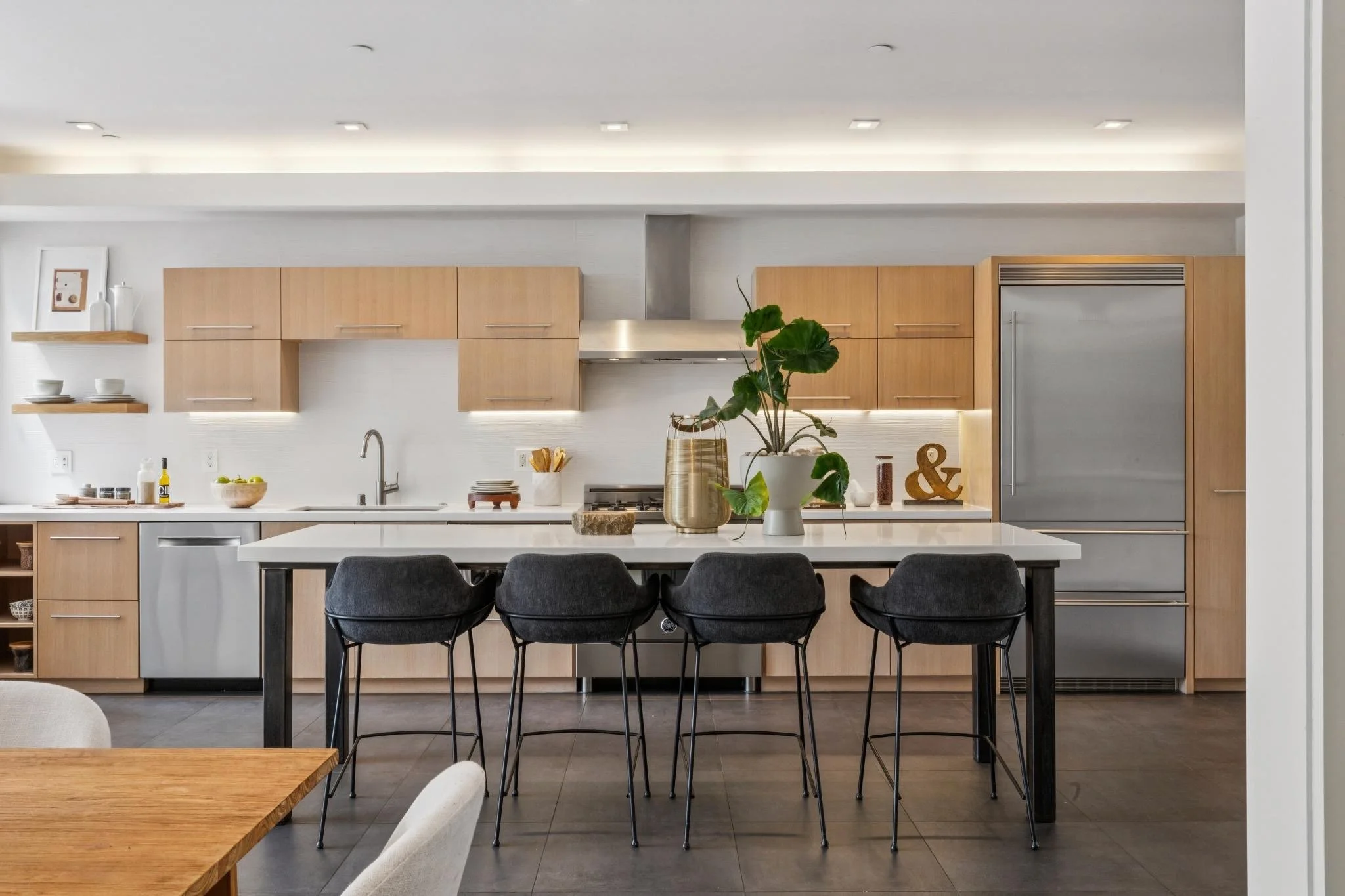 Modern kitchen with light wood cabinets, a white countertop, a large potted plant, bar stools at a kitchen island, and minimal decorative items.
