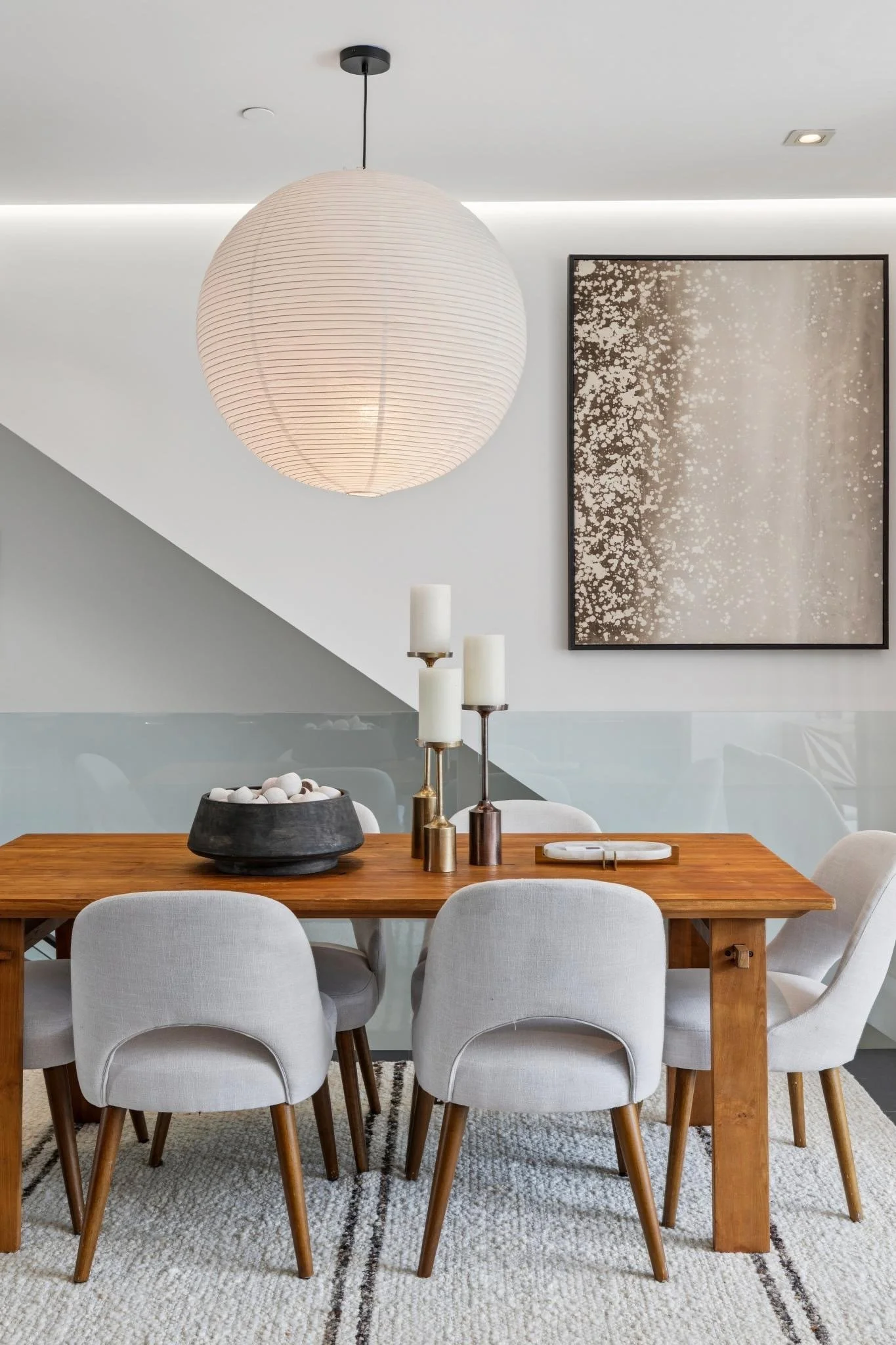 A modern dining room with a wooden table and eight white chairs, a large paper lantern hanging from the ceiling, three white candles in brass holders and a black bowl with white decorative objects on the table. There is a large abstract painting on t