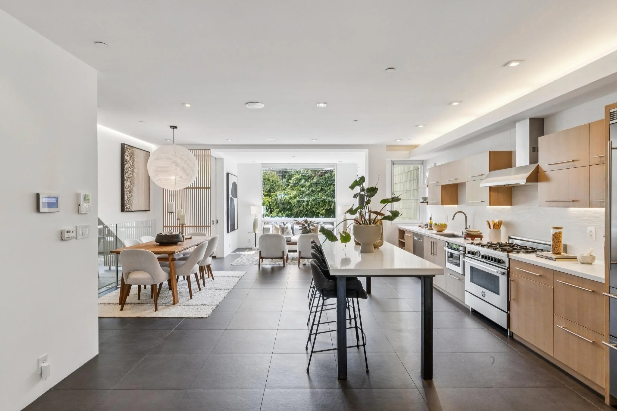 Open-concept modern kitchen and dining area with white walls, large windows, light wood cabinets, black countertops, and a dining table with chairs.