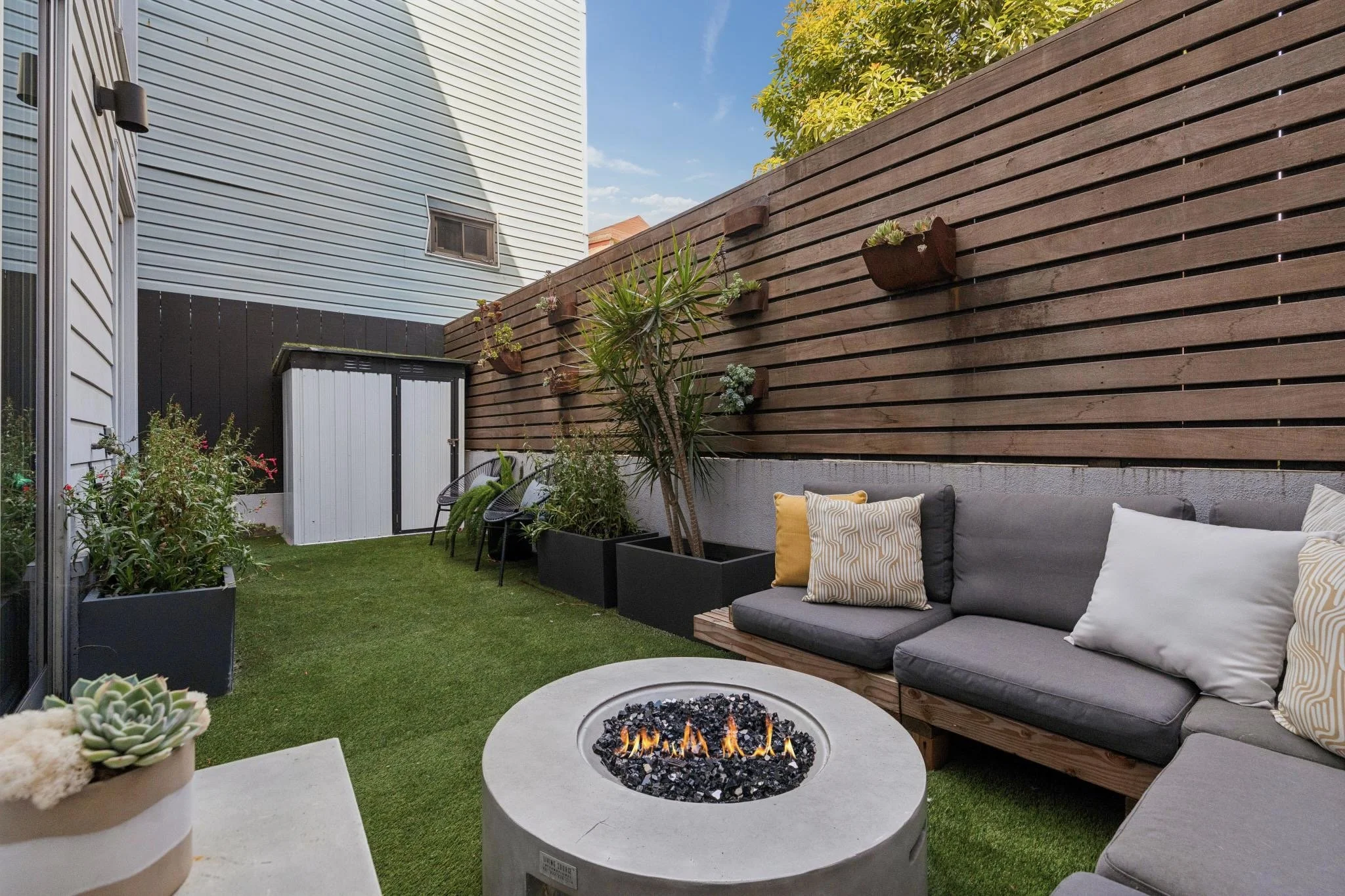 A cozy outdoor patio with a fire pit, a gray sectional sofa with pillows, potted succulents, and a wooden privacy fence with hanging planters. There is a small shed and a grassy area under a blue sky.