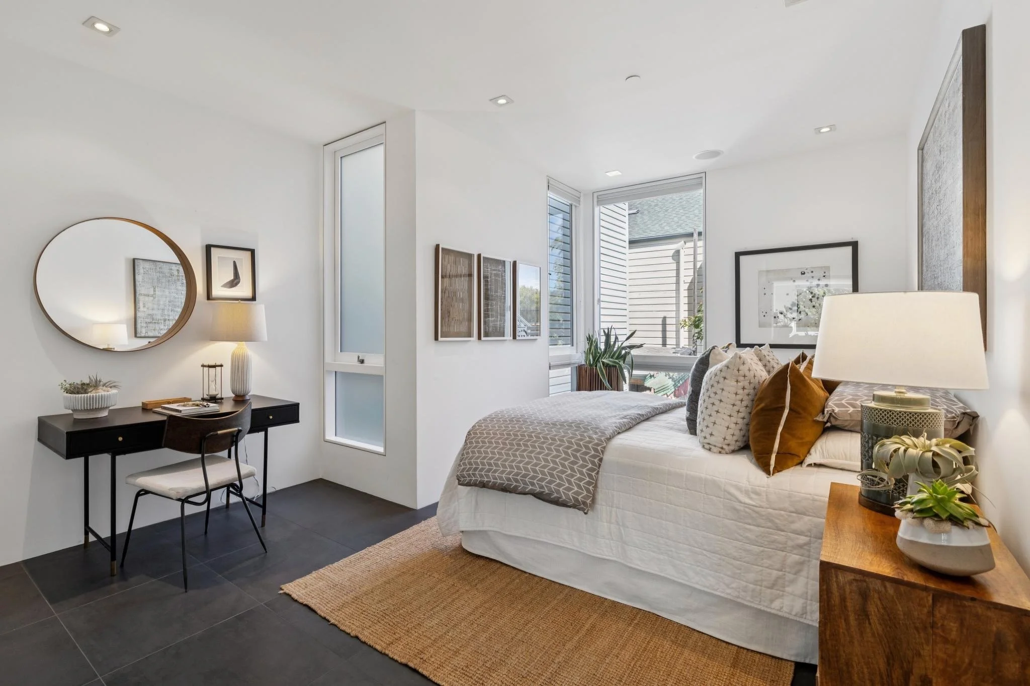 A modern bedroom with a white bed, pillows, and a blanket. There are large windows letting in natural light, framed photos on the wall, a small black desk with a chair, and decorative plants. The room has white walls with artwork and a tan rug on dar