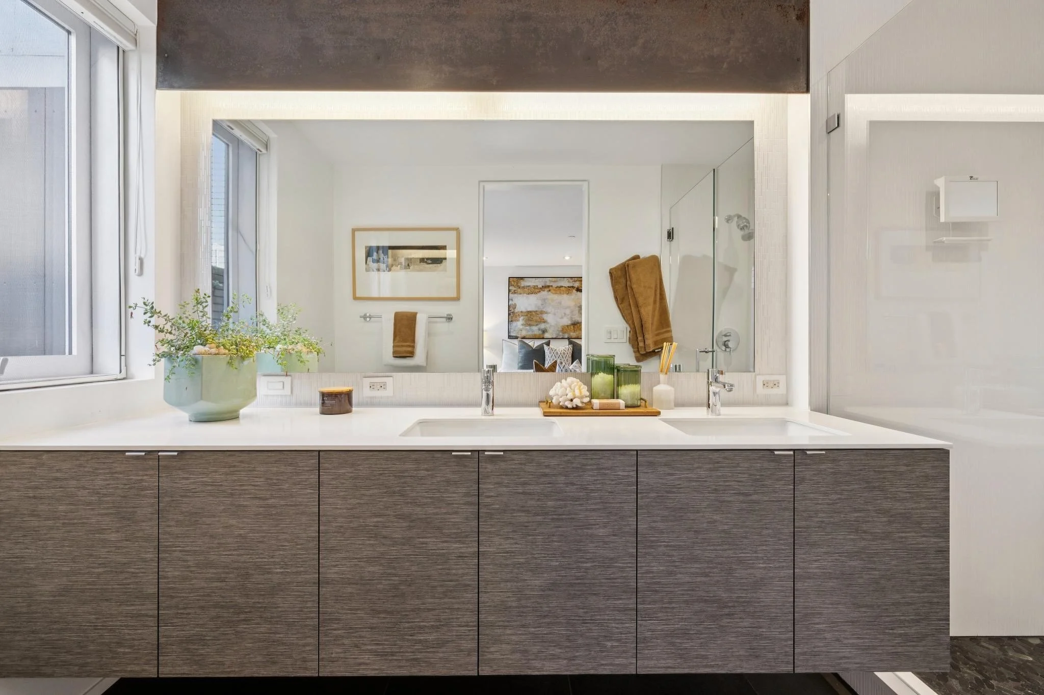 Modern bathroom with a wide mirror, white countertop, plant in a pot, and various toiletries, with a window on the left and a textured wall at the top.