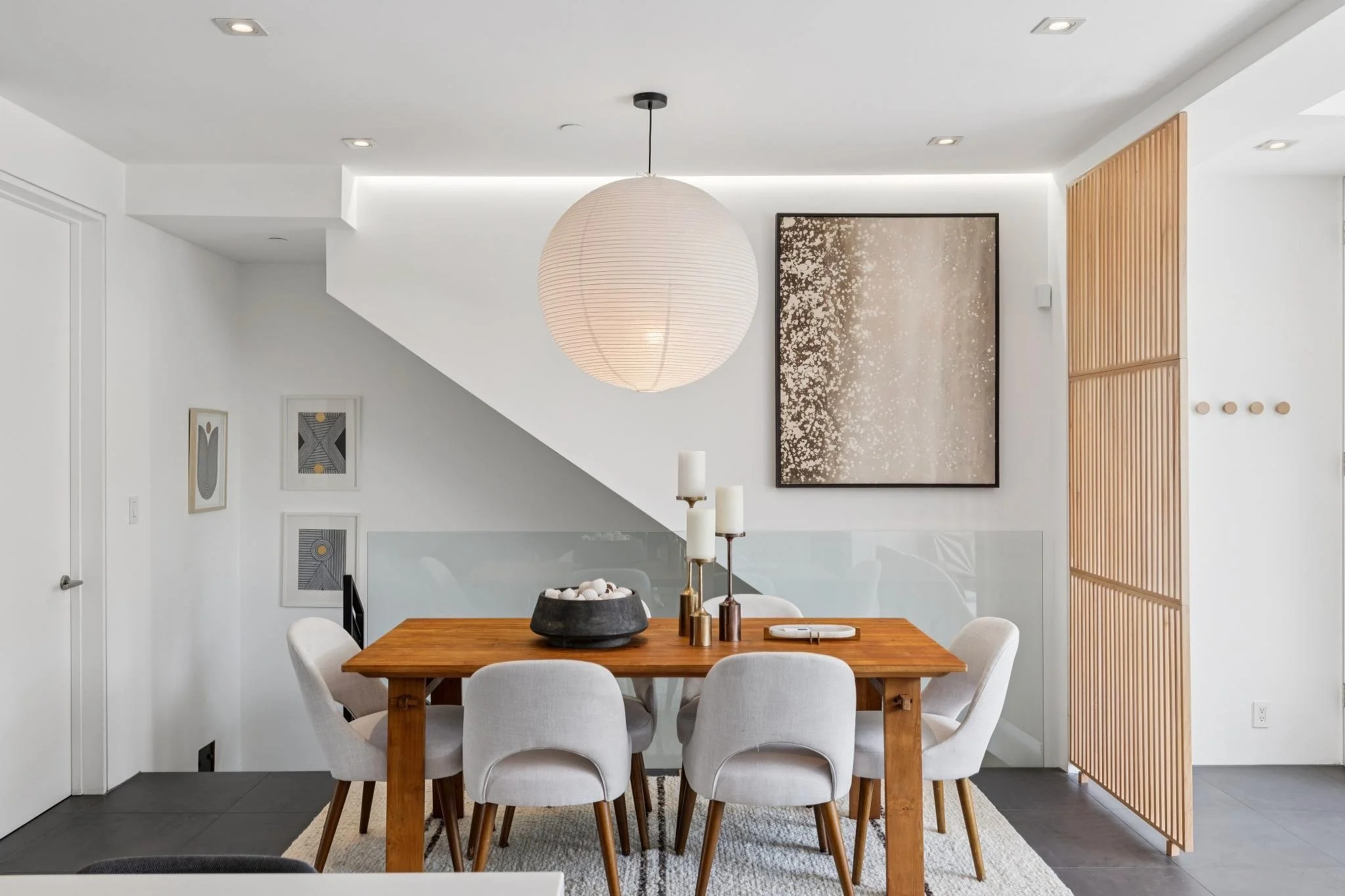 Modern dining room with wooden table, white chairs, large paper lantern pendant light, abstract wall art, candles, and wooden slatted partition.
