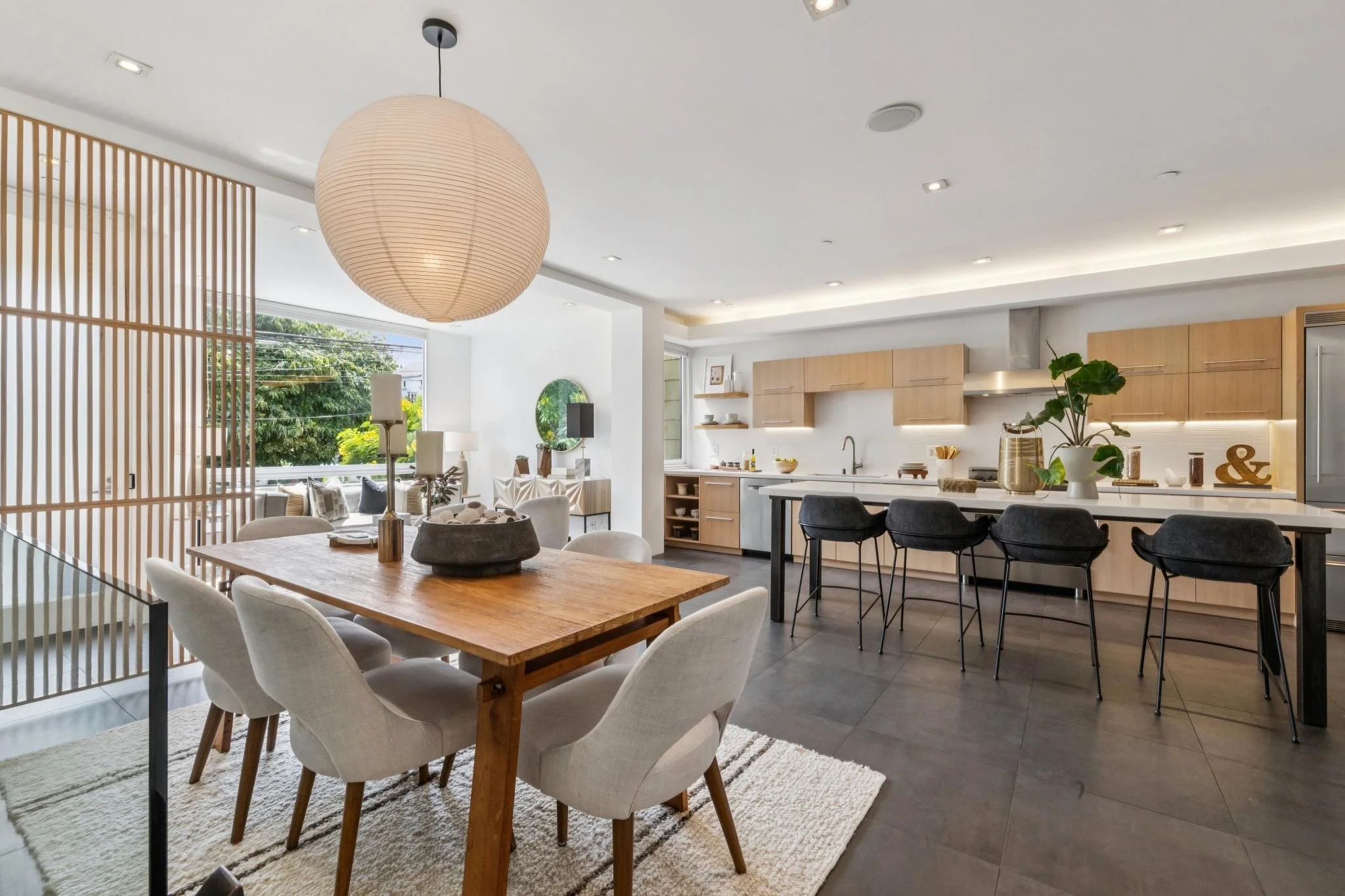 Open-concept kitchen and dining area with wooden dining table surrounded by white upholstered chairs, adjacent to a modern kitchen with light wood cabinetry, black barstools, and kitchen appliances. Large windows let in natural light, with a hanging 