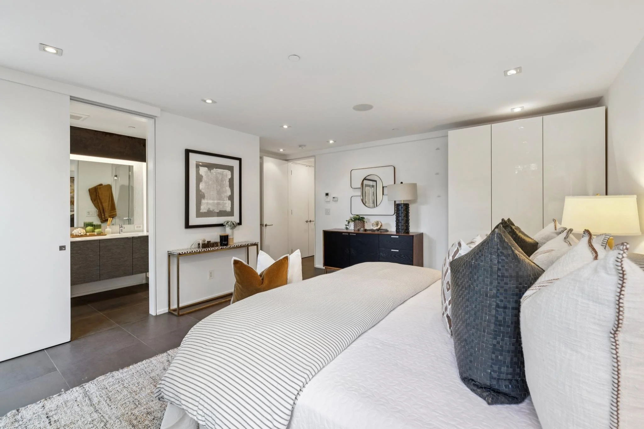 Modern bedroom with a neatly made bed featuring white and patterned pillows, a nightstand with a lamp, and wall decor. An open doorway leads to a bathroom with a mirror and towel rack.
