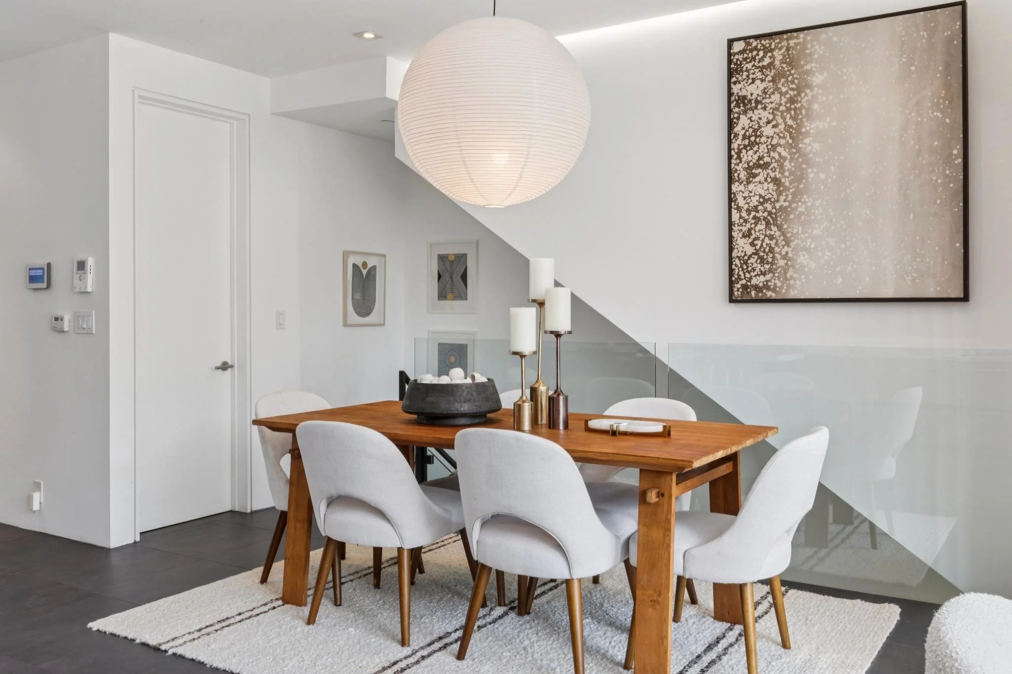 A modern dining room with a wooden table surrounded by white cushioned chairs, a large white paper lantern pendant light, wall art, and decorative candles on the table.