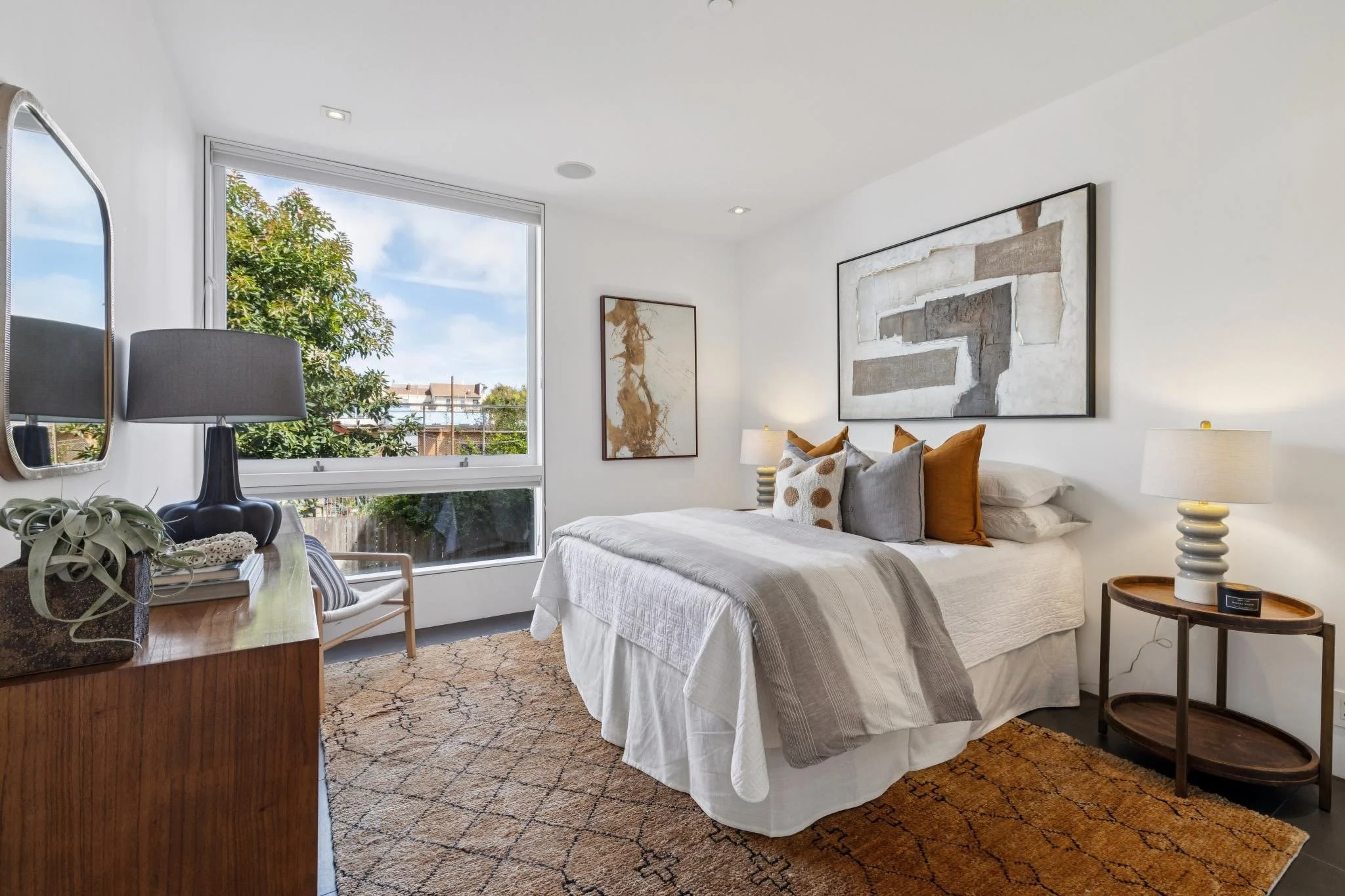 A bright bedroom with a large window showing trees and houses outside. The bed has white bedding with decorative pillows in shades of brown, gray, and white. There are two bedside lamps with white shades. On the left, a wooden table holds a plant, a 