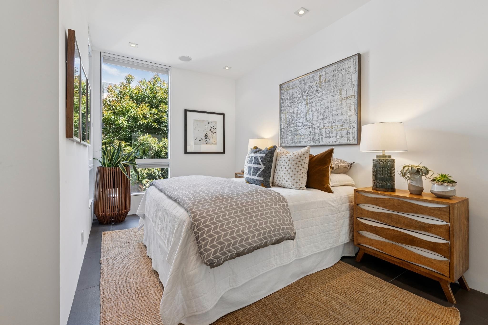 Modern bedroom with white walls, large window with greenery outside, artwork on walls, bed with patterned pillows and throw, wooden nightstand with lamp and decorative items.