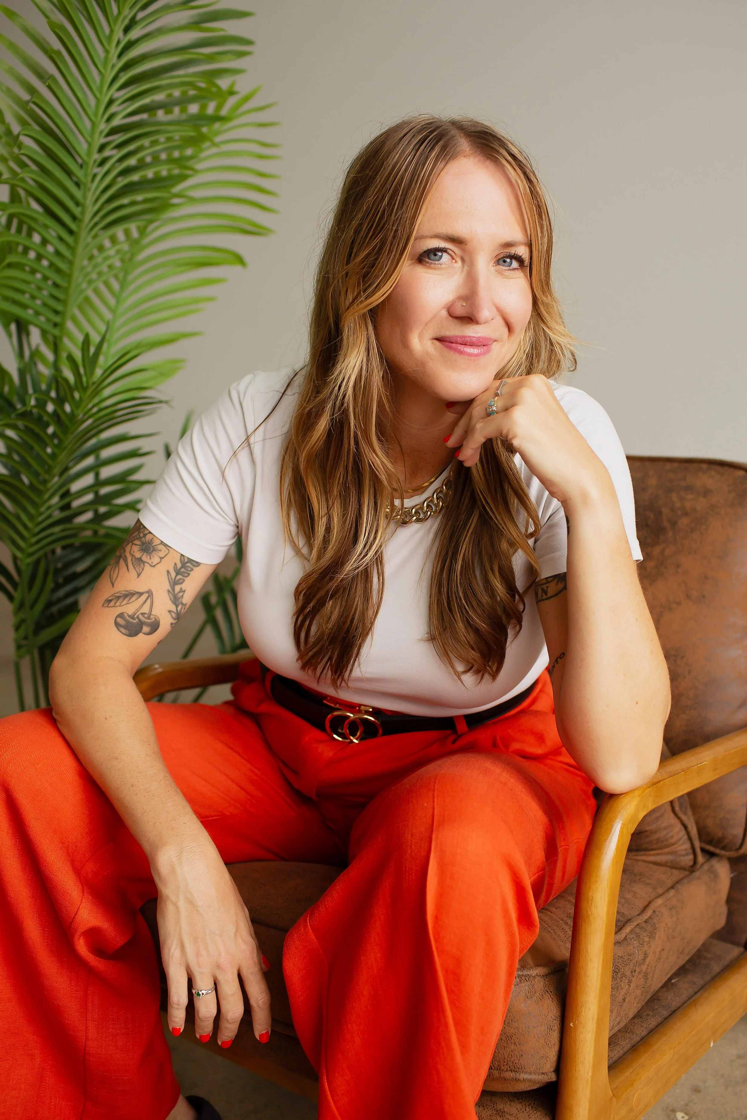 A woman with long, wavy, reddish-brown hair, sitting on a brown couch. She is wearing a white t-shirt, red pants, a gold chain necklace, and a ring. She has tattoos on her left arm and is resting her chin on her right hand, smiling slightly. There is a green plant on her left side.