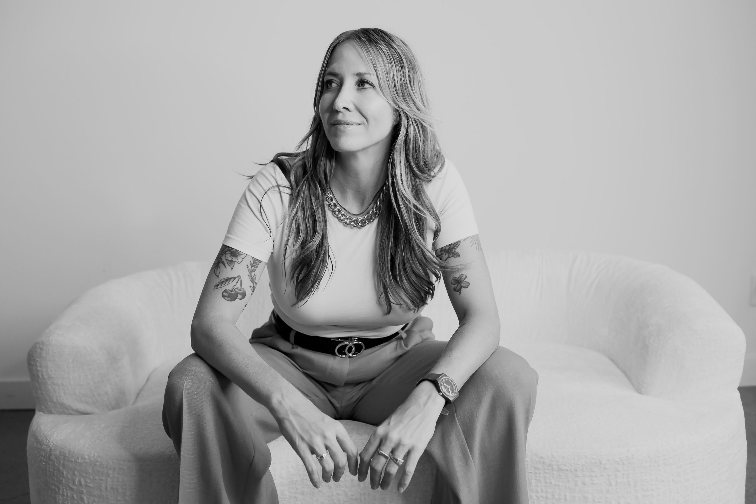 A black and white photograph of a woman sitting on a round cushioned seat. She has long wavy hair, several tattoos on her arms, and is wearing a white t-shirt, beige pants, a chunky necklace, a watch, and rings. She is smiling softly and looking to her left.