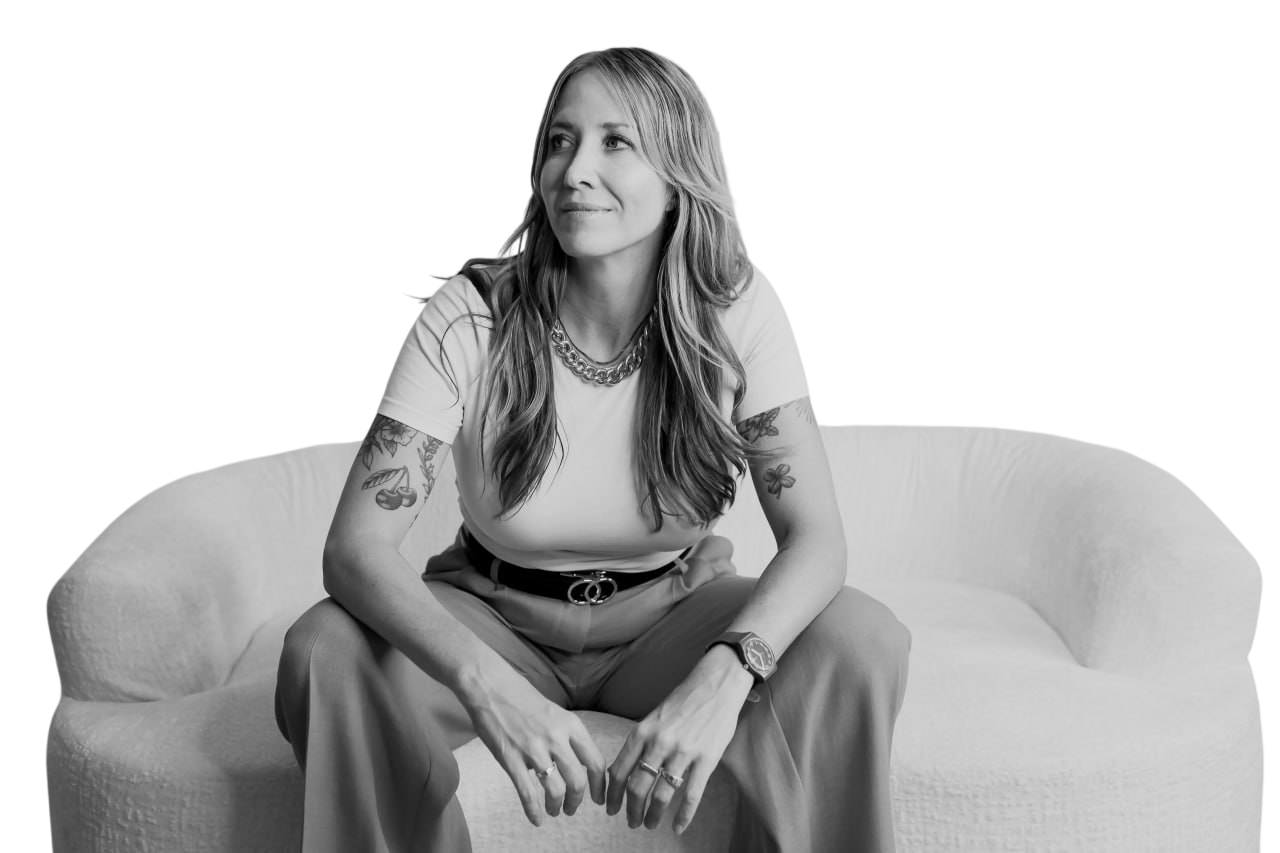 A woman with long hair, tattoos on her arms, wearing a white t-shirt, trousers, a necklace, a watch, and rings, sitting on a curved cushioned sofa.