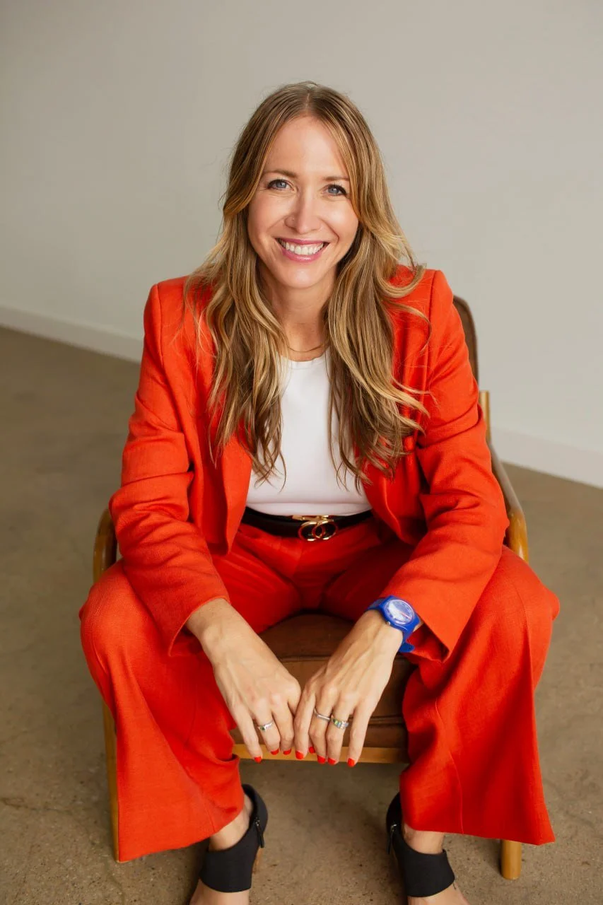A woman with long hair wearing a red suit, white top, and black shoes, sitting on a wooden chair, smiling at the camera.