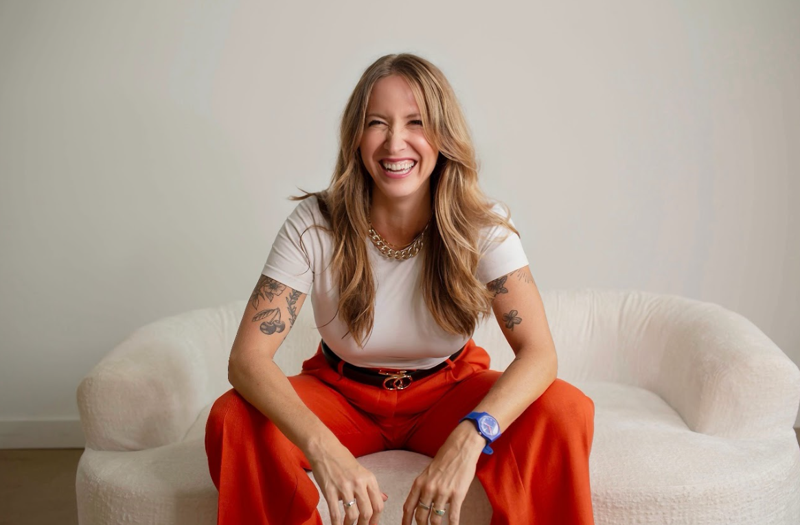 A woman with long blonde hair, smiling and sitting on a white sofa, wearing an off-white T-shirt, red pants, a necklace, and a blue watch.