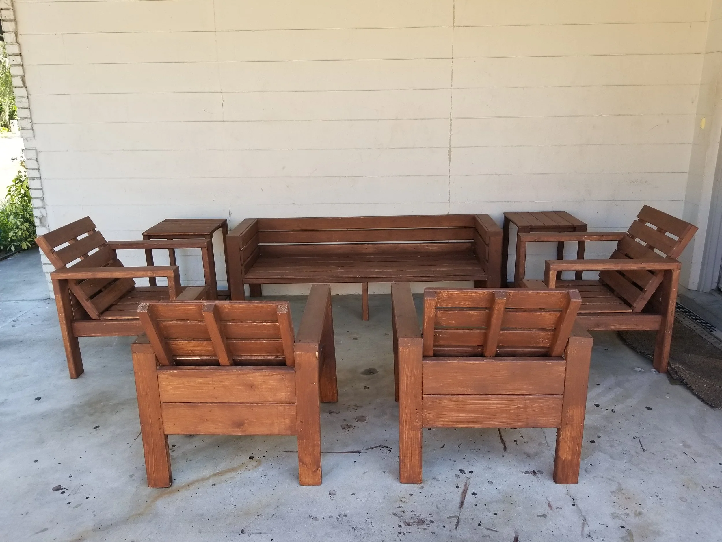 A set of wooden outdoor furniture including a sofa, two armchairs, and two side tables, arranged on a concrete patio against a light-colored wall.