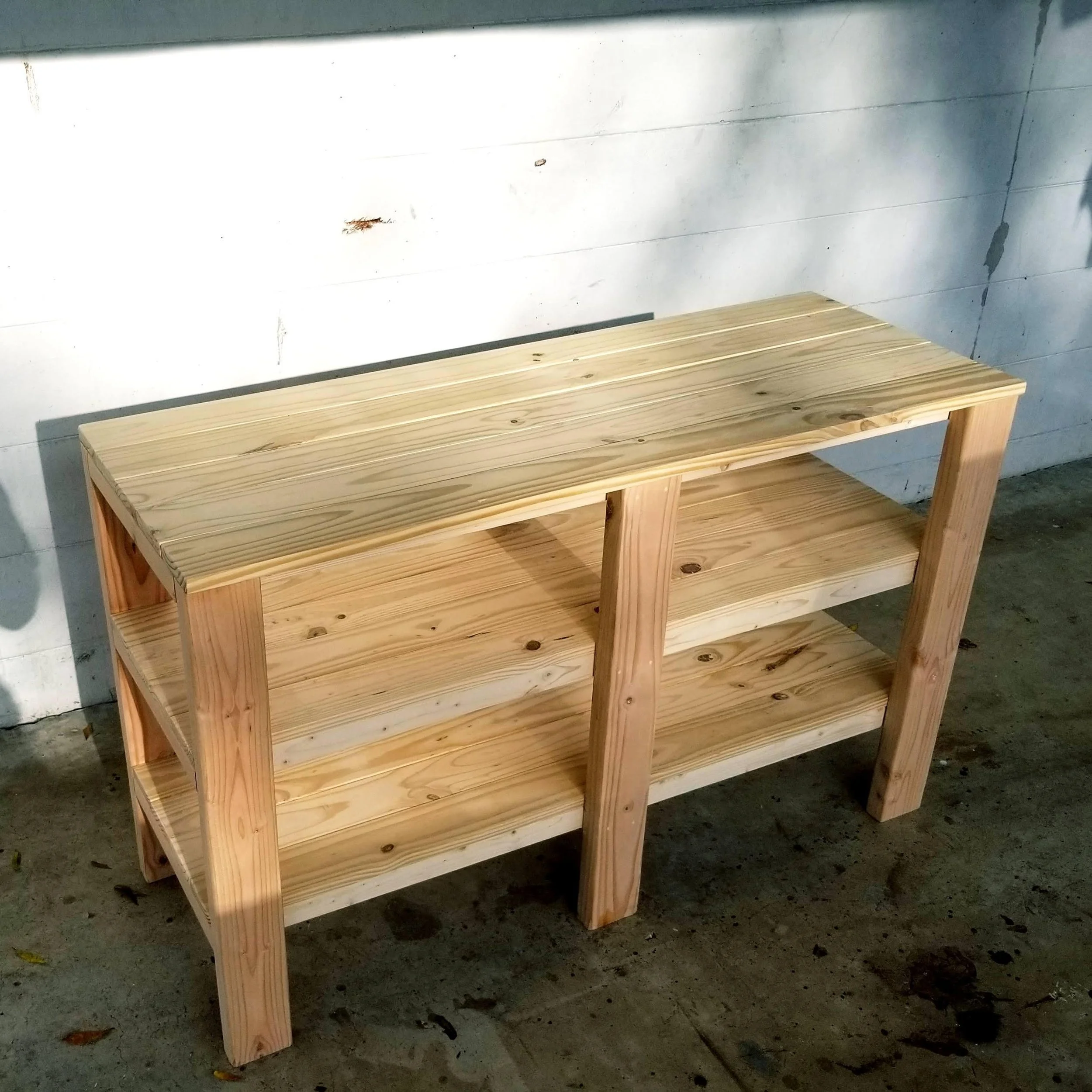 A wooden bench with four slatted shelves in a gray basement or garage, with a white wall behind it and a concrete floor with some stains and leaves.
