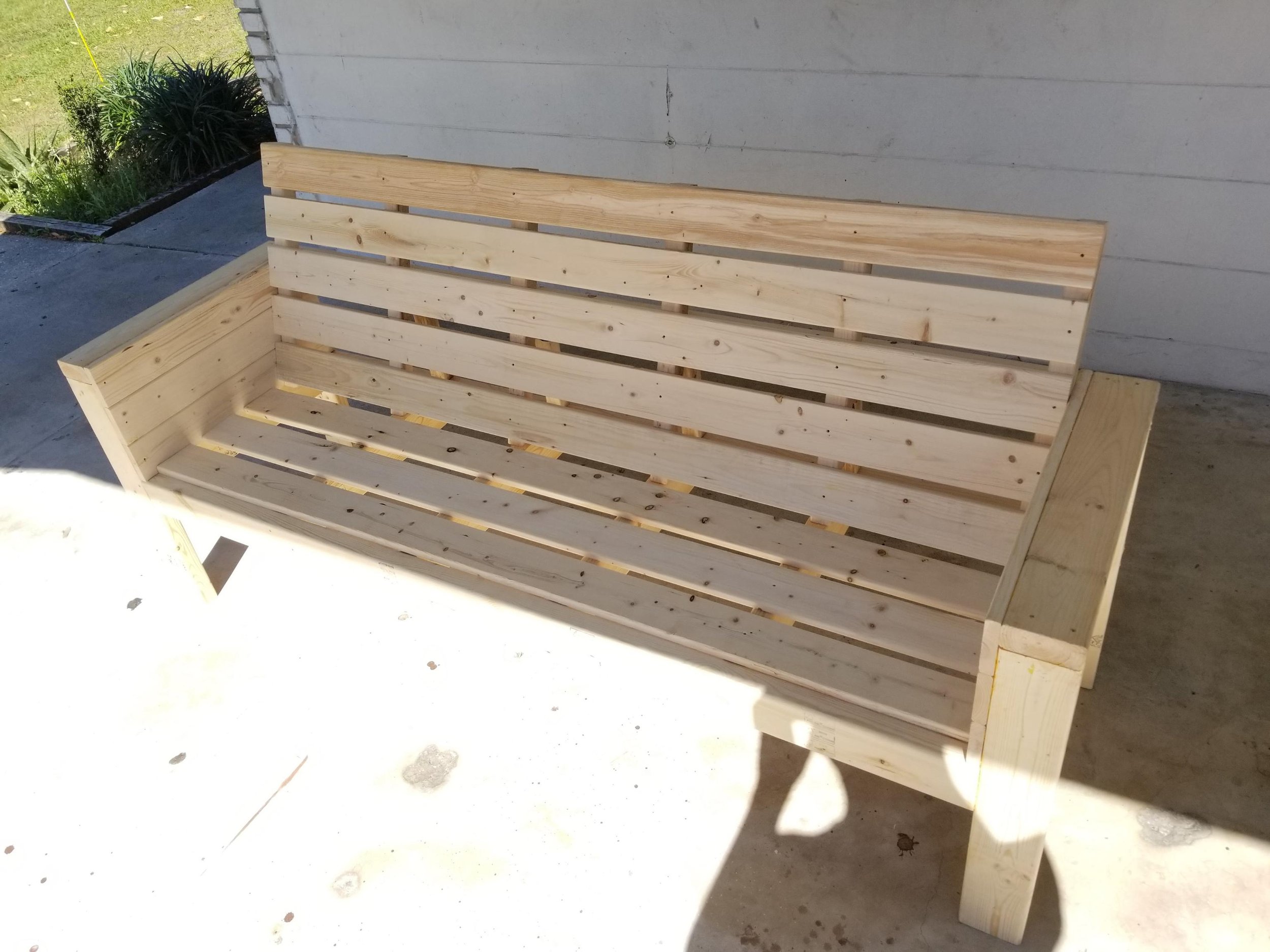 A wooden bench with a slatted backrest and seat, positioned outside near a house wall, with some plants visible in the background.