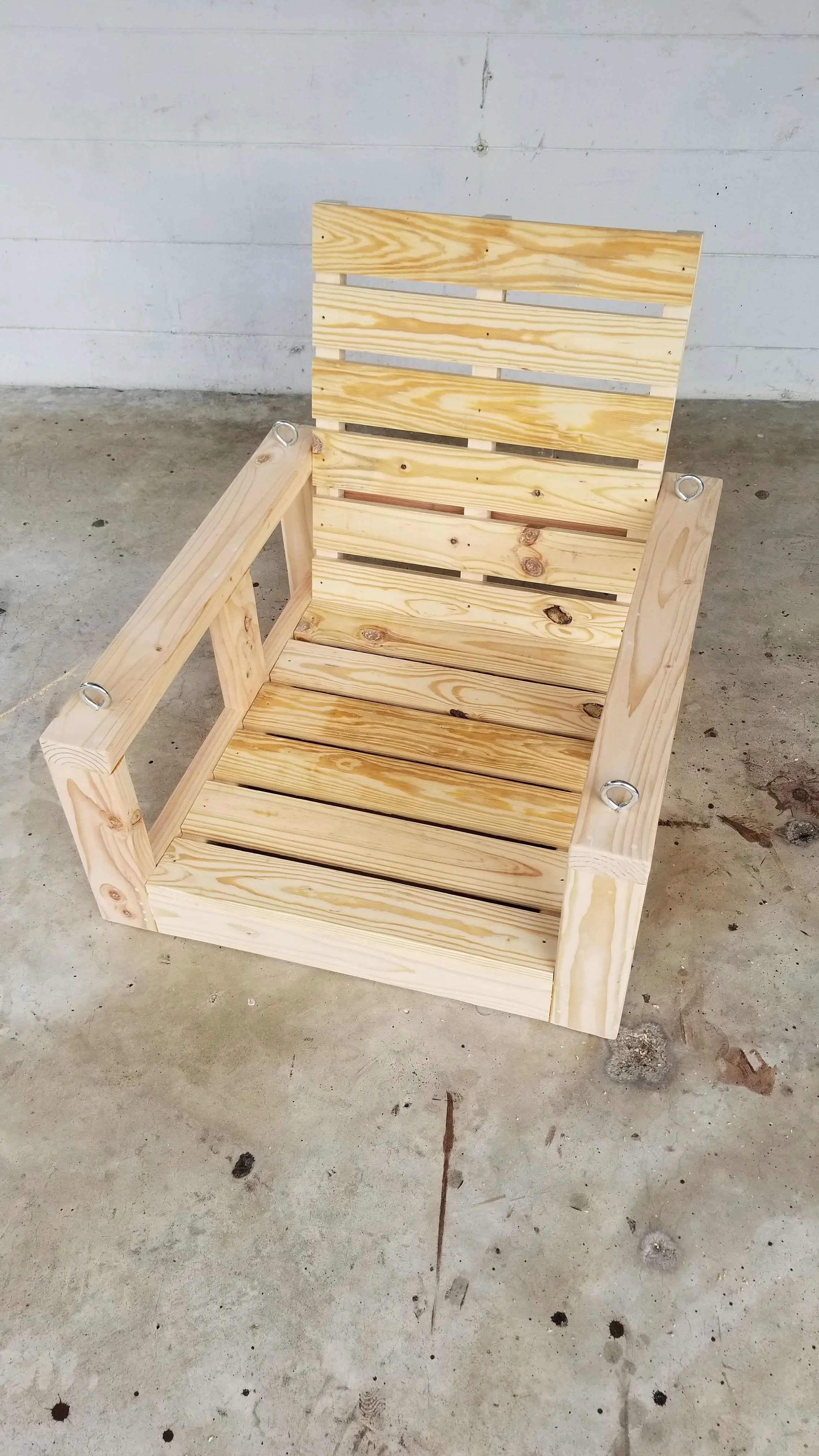 A wooden Adirondack-style chair made of unfinished light wood, situated on a concrete floor against a white wall.