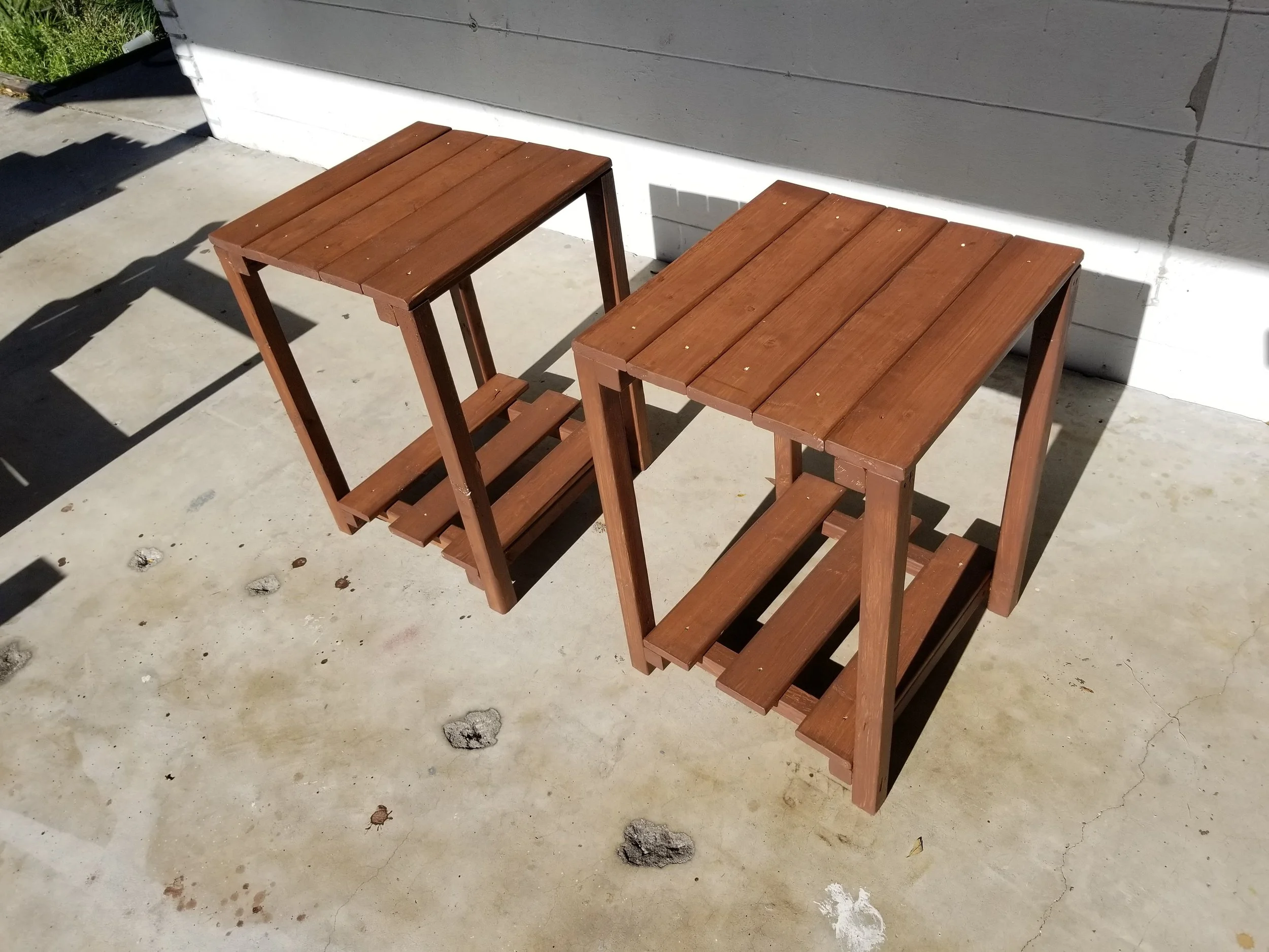 Two wooden tables on a concrete ground outside, with a gray painted wall in the background.
