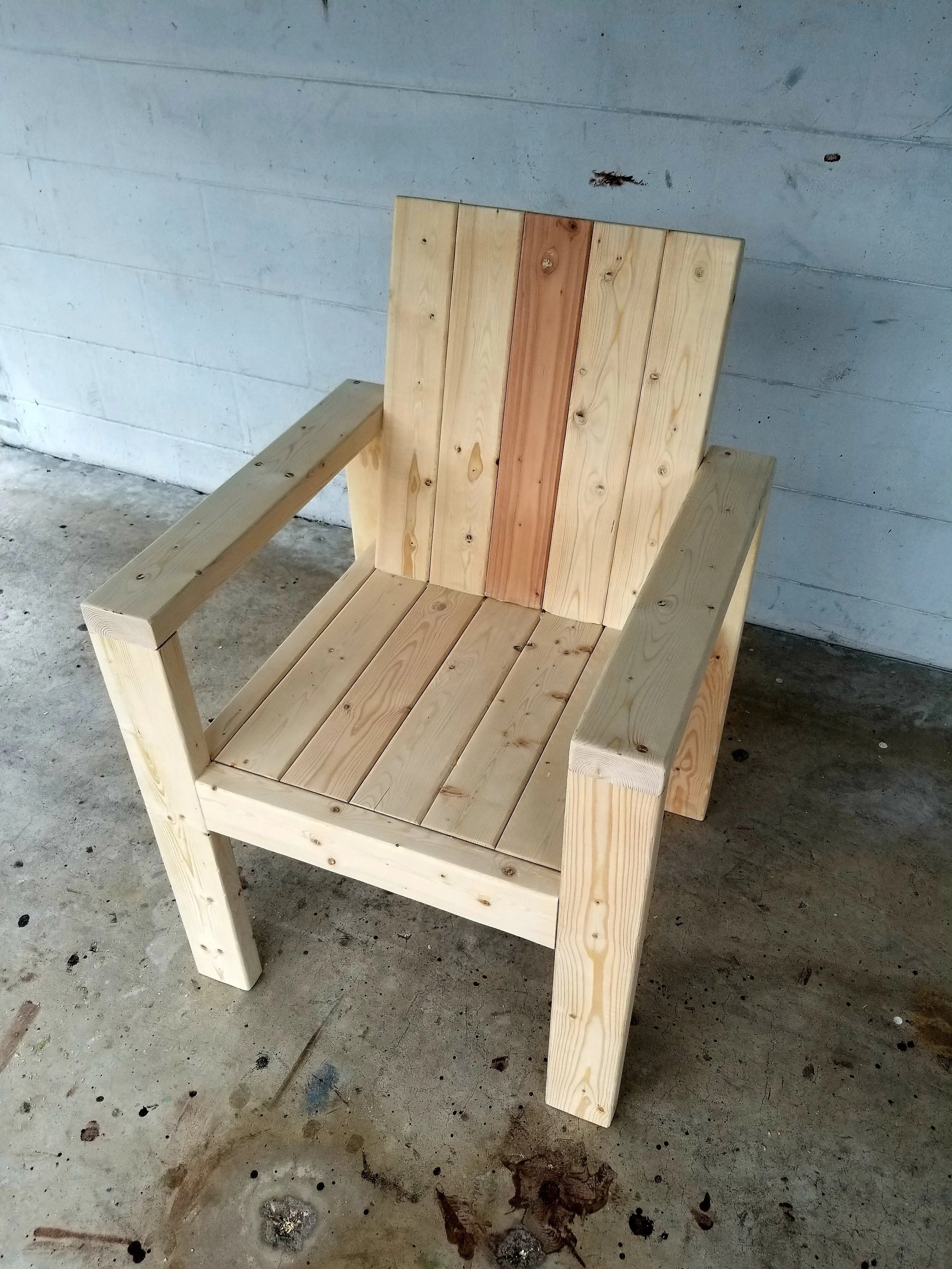 A wooden outdoor chair with a slatted backrest and seat, positioned on a concrete floor against a painted wall.