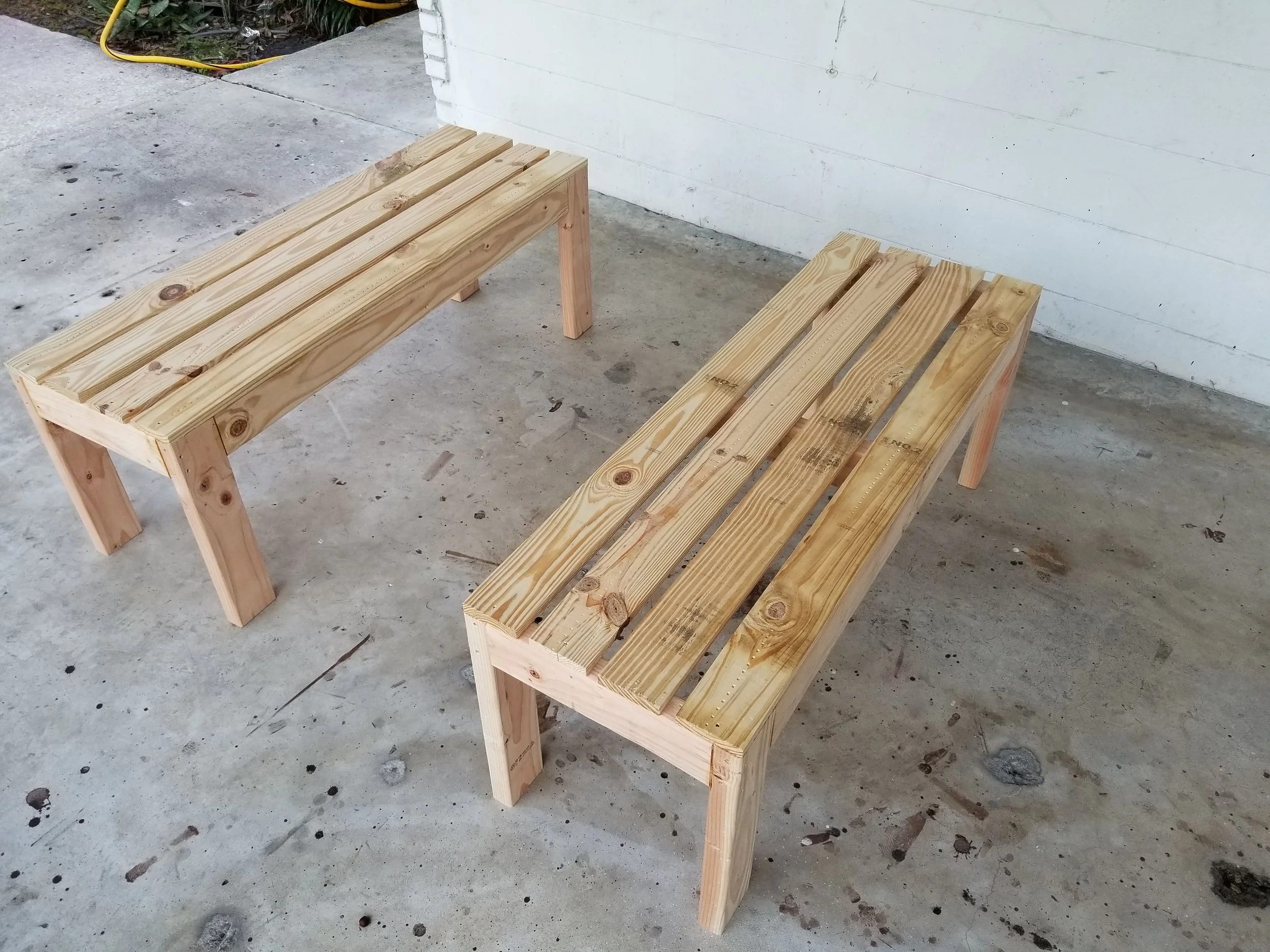 Two wooden benches made of unfinished light-colored wood on a concrete porch, with a white wall and some plants in the background.