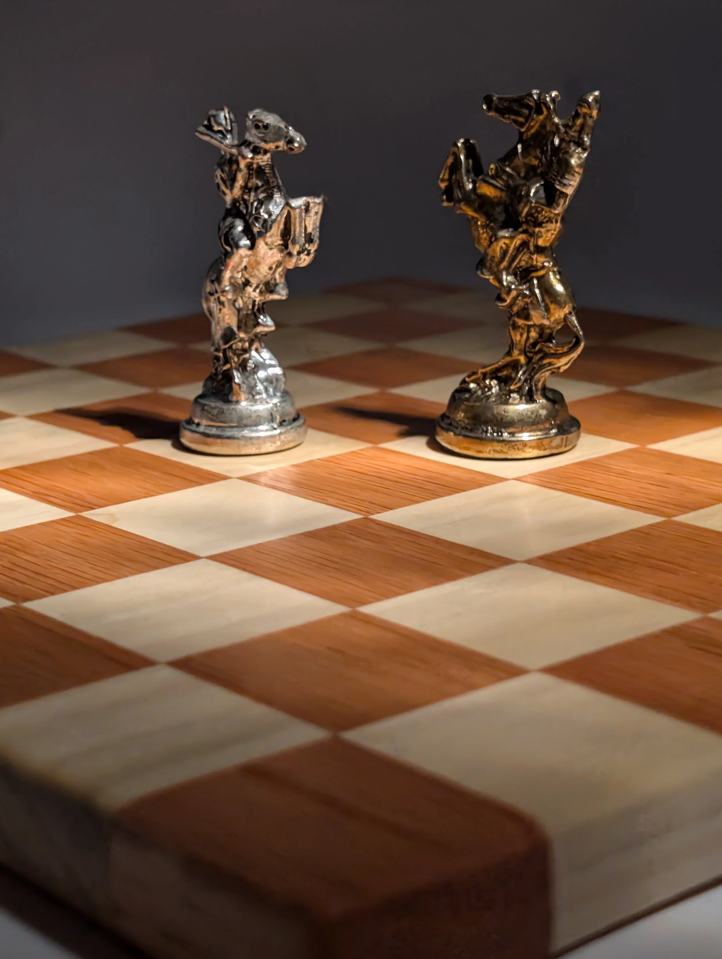 Two metallic chess king pieces on a wooden chessboard with a checked pattern, against a dark background.