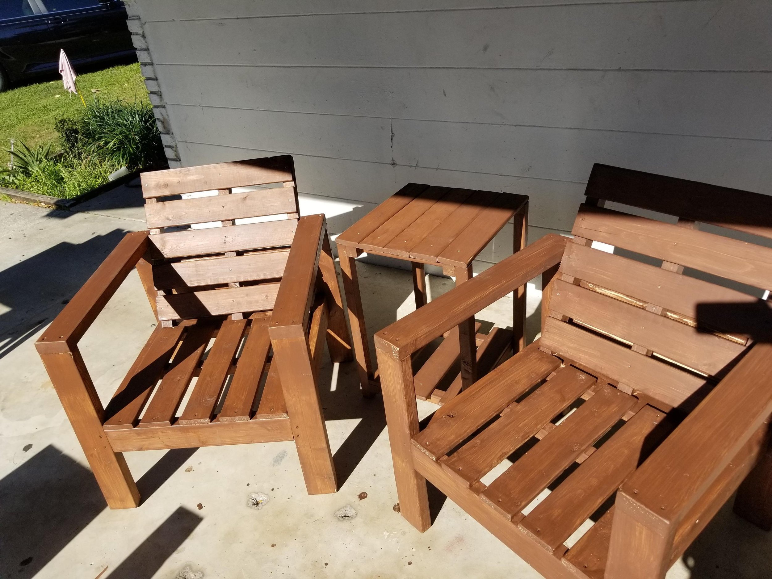 Outdoor patio area with wooden chairs and tables, some chairs are stacked. There is a gray wall in the background, and a small garden with green plants and an umbrella can be seen on the left.