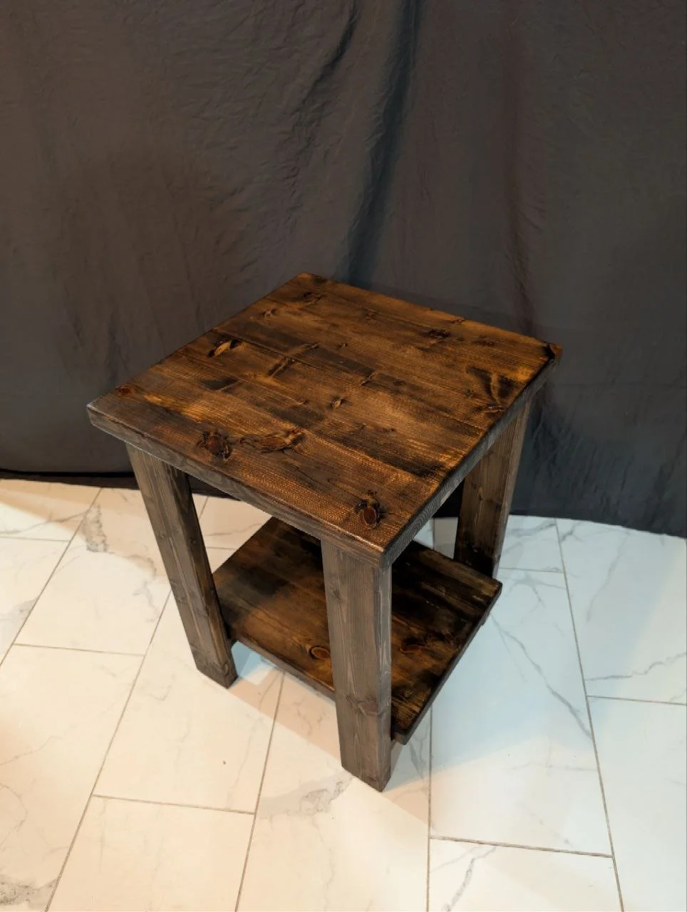 Small wooden nightstand with two shelves, dark wood finish, on a white marble floor with gray veining.
