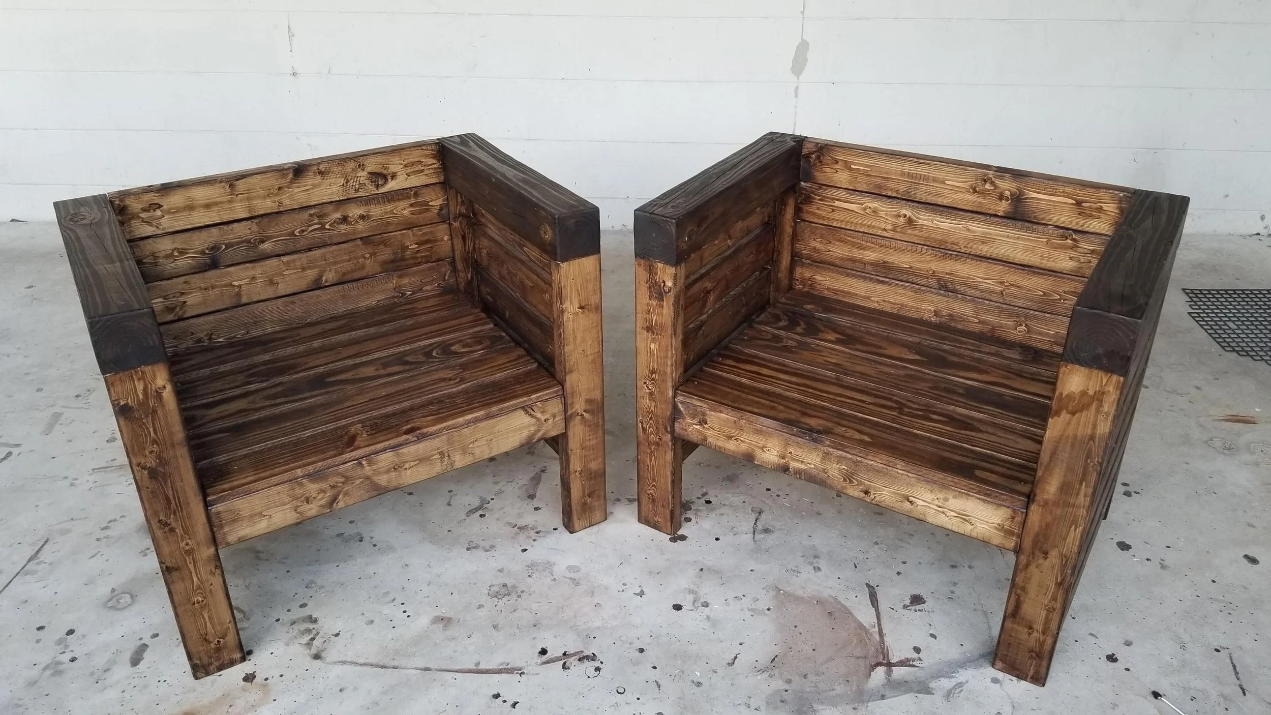 Two wooden benches with slatted backs and seats placed side by side on a concrete floor in front of a plain white wall.