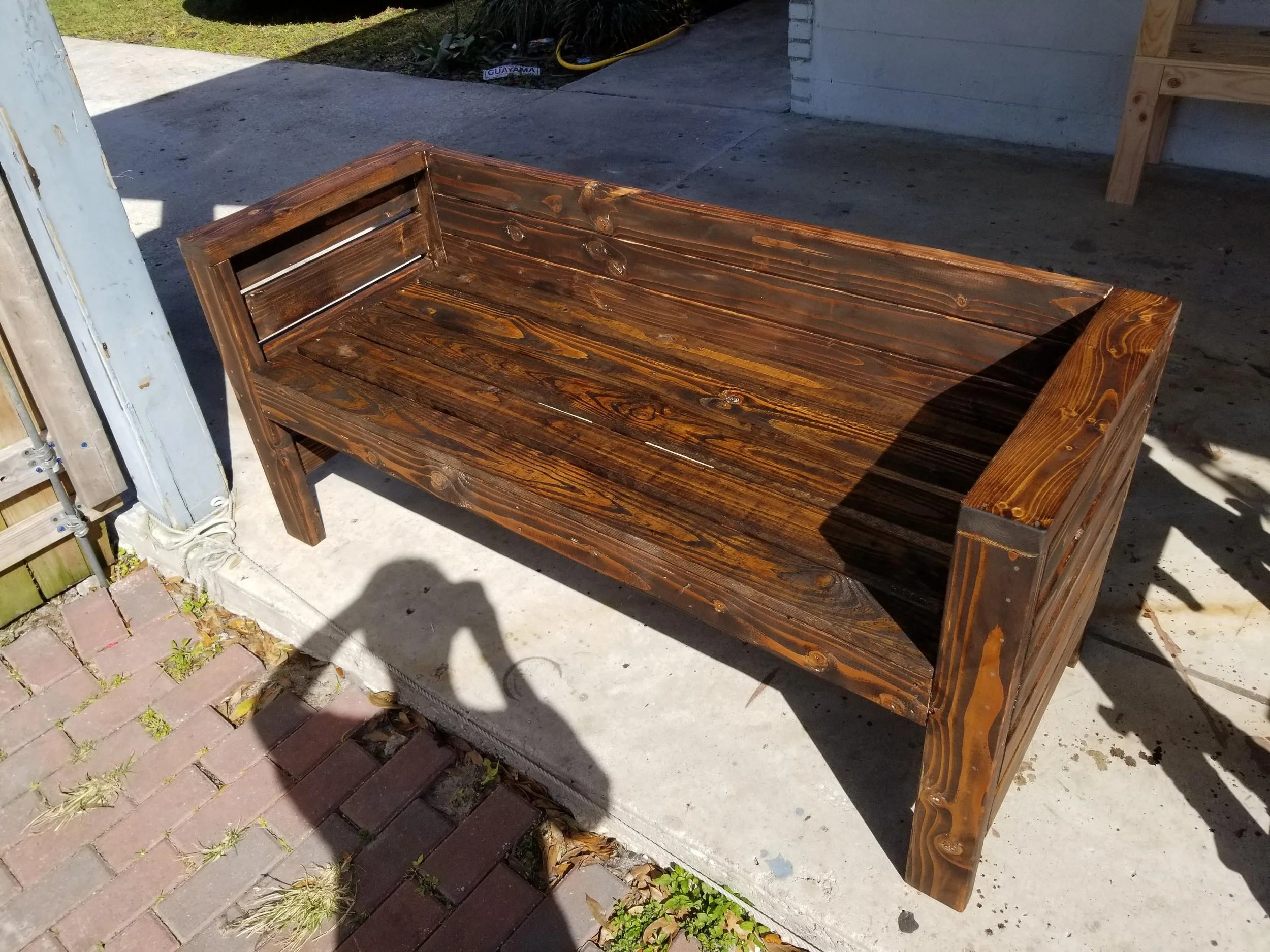 A wooden outdoor bench sits on a concrete surface near a brick sidewalk. The bench is made of stained wood planks with armrests and a slatted backrest. The background shows a garden area with grass and other wooden structures.