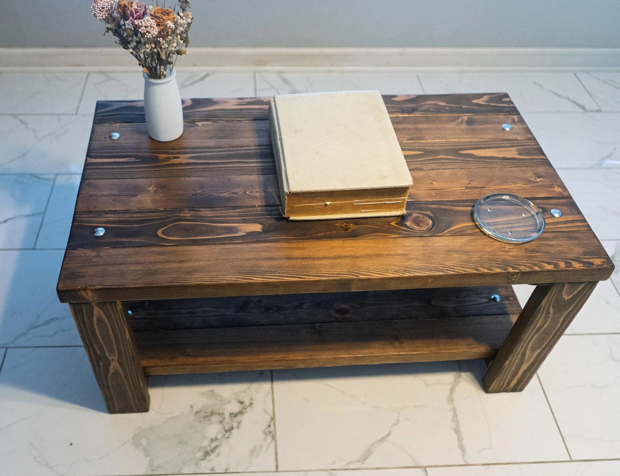 A dark wooden table with a white book, a white vase of dried flowers, a glass of water, and metal knobs on the top surface, situated on a white tile floor.