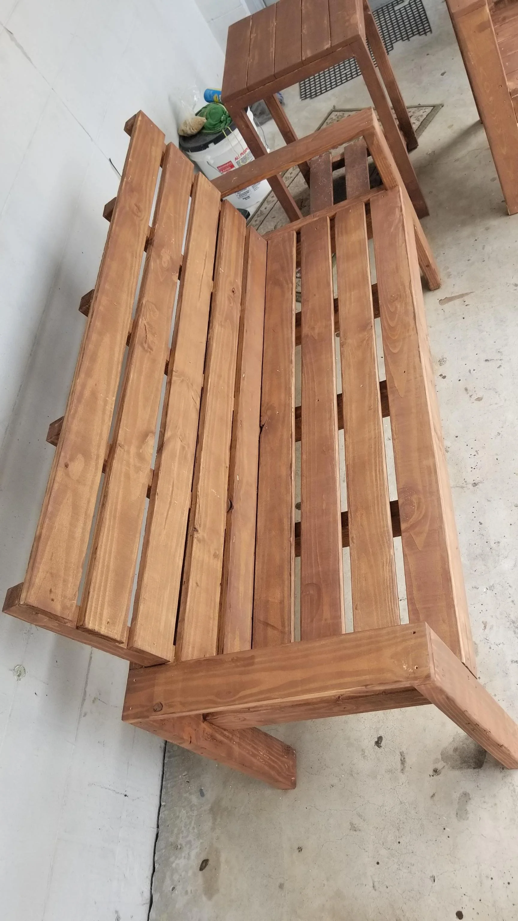 A wooden bench with a slatted seat and backrest, positioned on a concrete floor next to a wall. In the background, there are various items including a bucket, a container, and some tools.