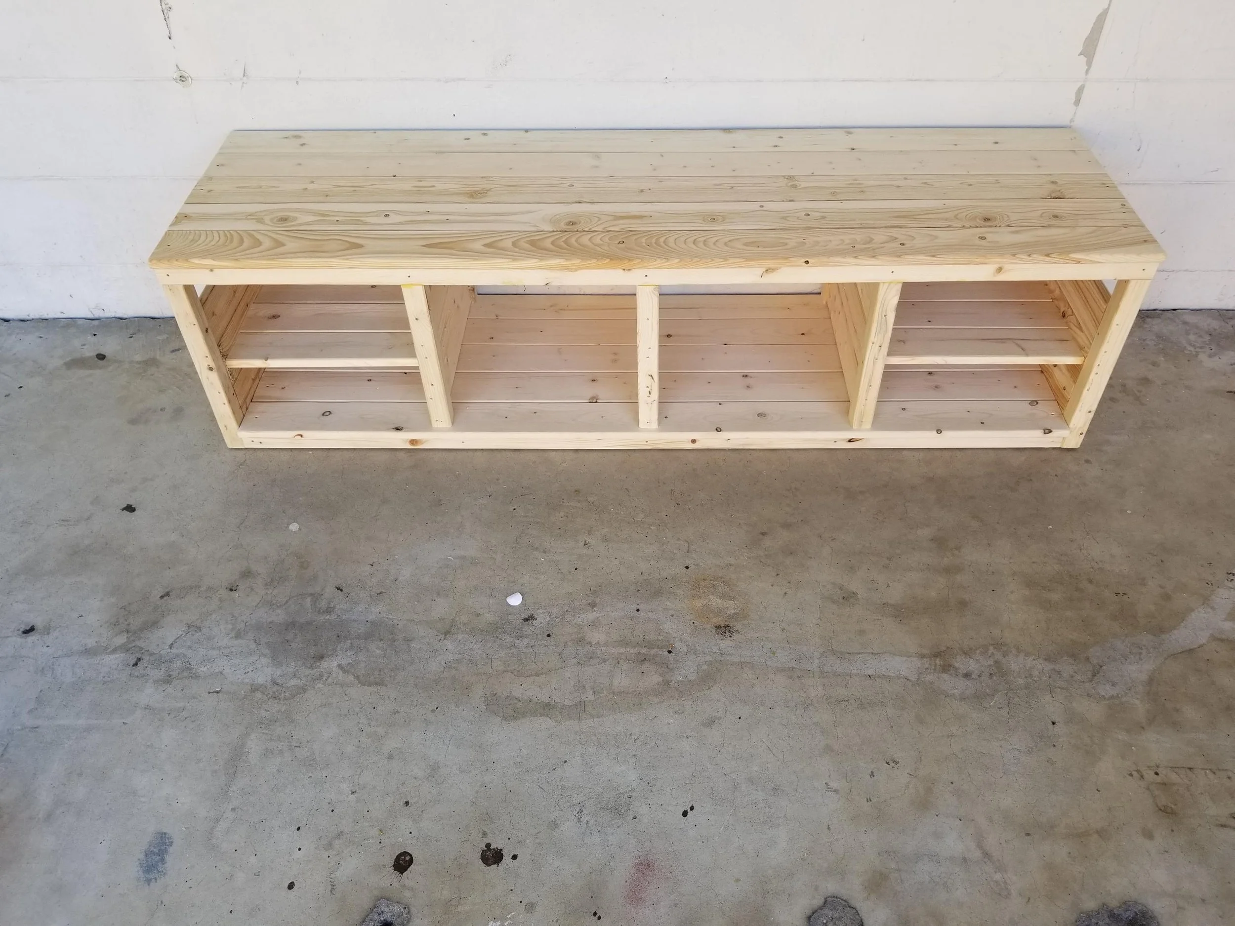 Unfinished wooden bench with three compartments on a concrete floor against a white wall.