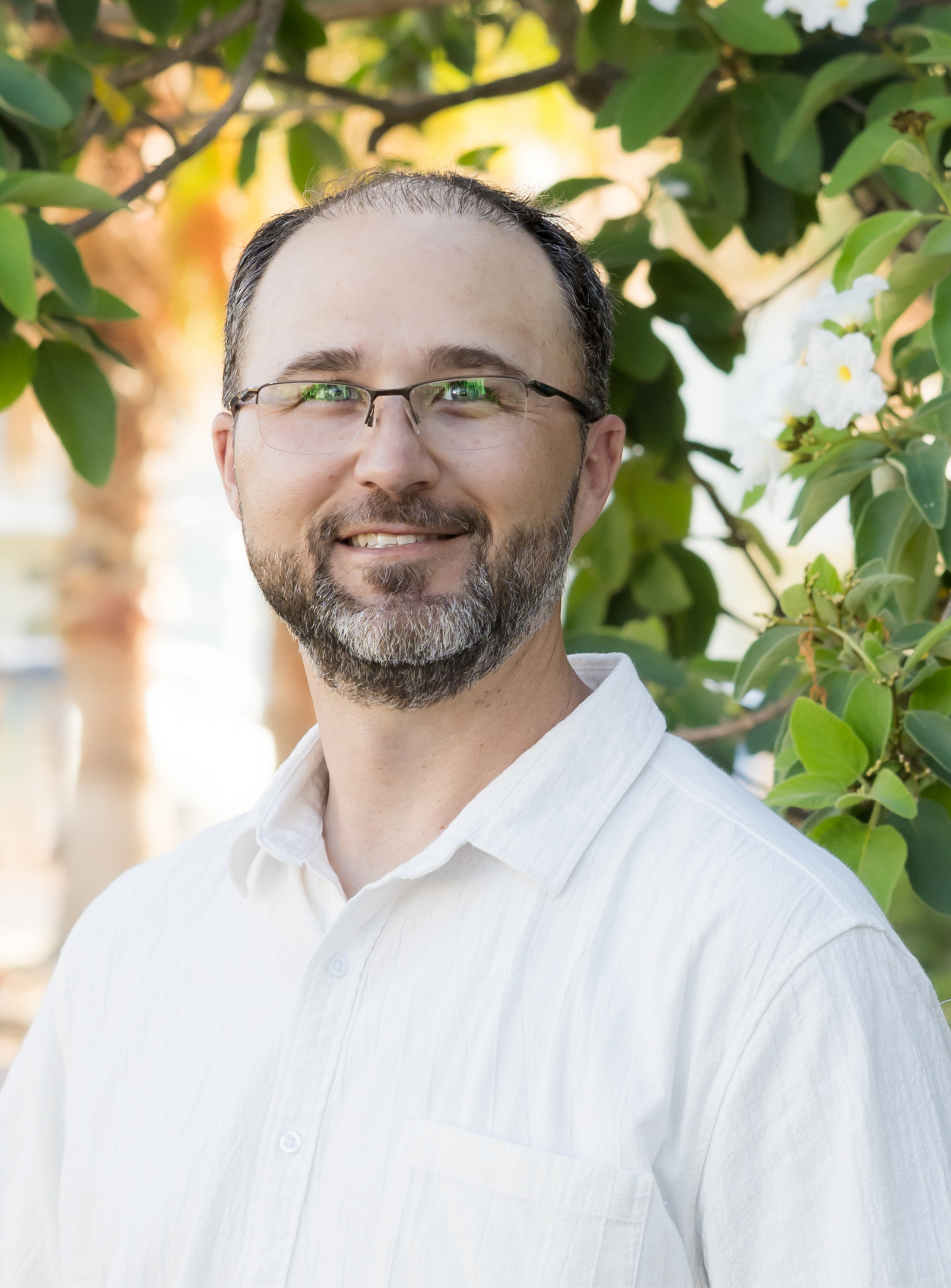 A man with glasses, a beard, and short brown hair, smiling, wearing a white shirt, standing outdoors amidst green foliage and white flowers.