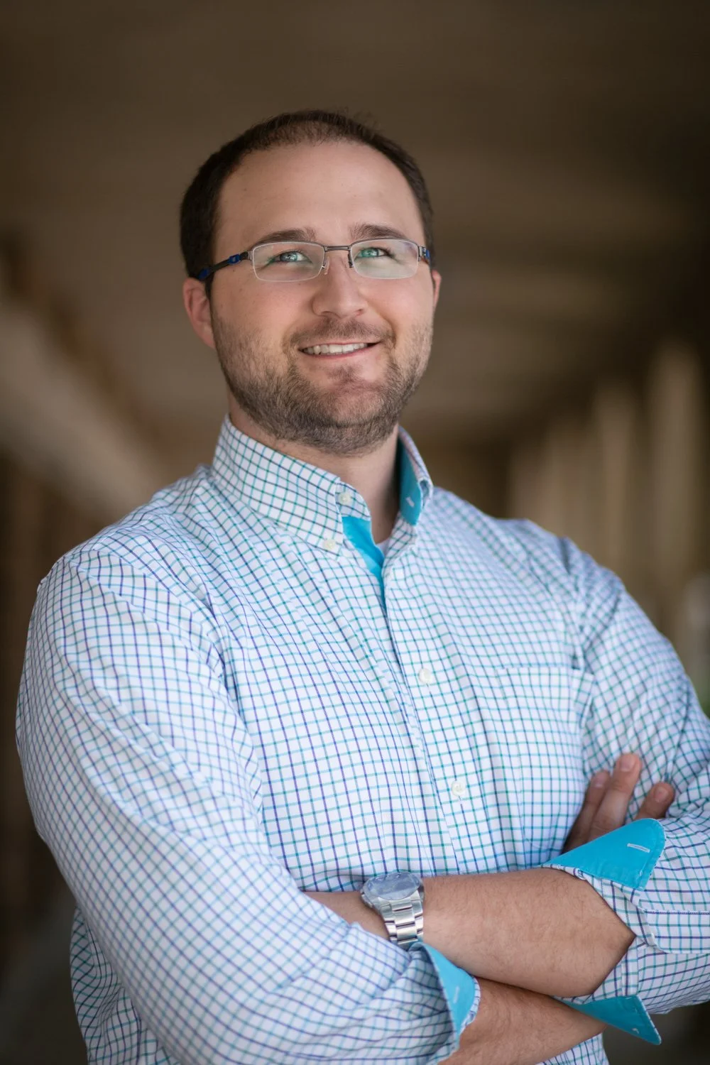 A man with glasses and a beard, wearing a checkered button-up shirt and a wristwatch, standing with arms crossed and smiling.