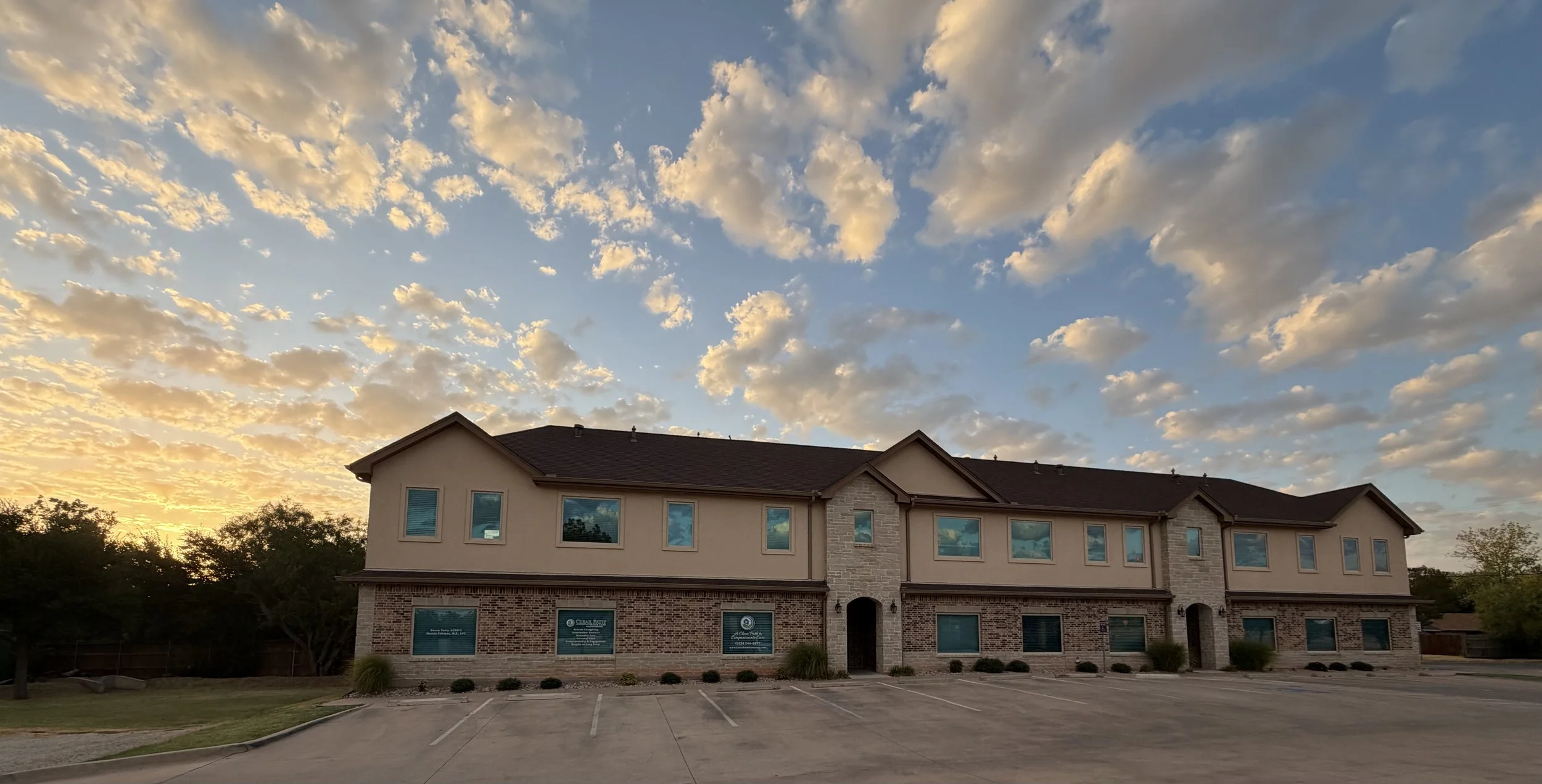 A two-story residential building with brick and beige exterior walls, multiple windows, and an arched doorway, set against a partly cloudy evening sky with the sun setting near the horizon.