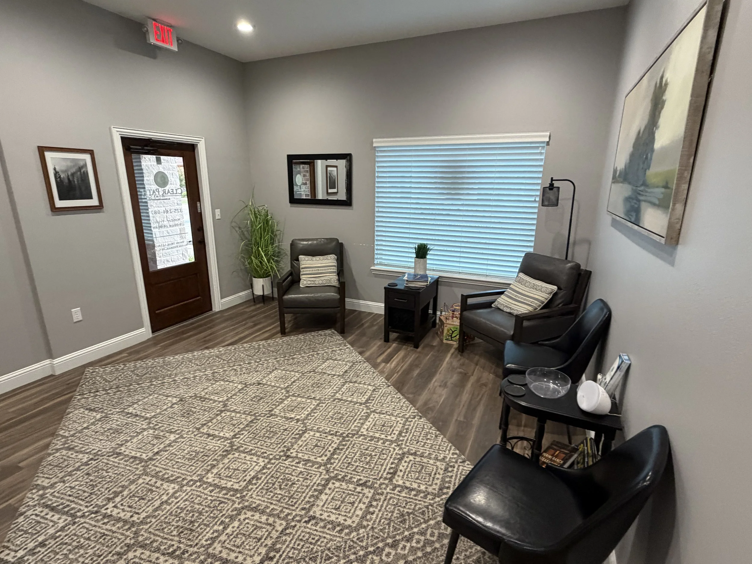 Waiting room with gray chairs, a black table with magazines, plants, framed pictures on gray walls, and closed blinds on a window.