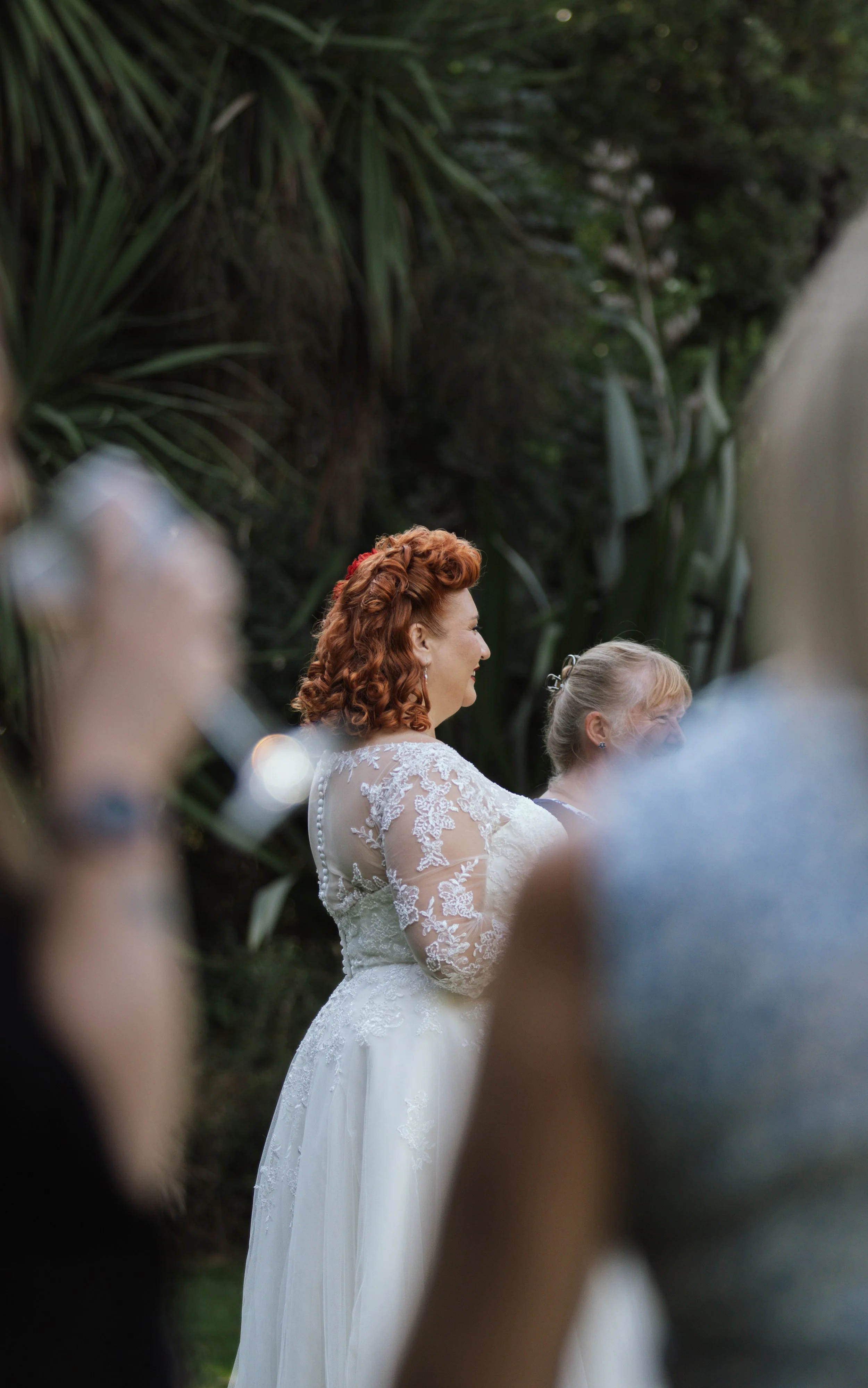A bride with red, curly hair wearing a white lace wedding gown, standing outdoors near lush green foliage. She is smiling and looking to the right, with a person next to her partially visible.
