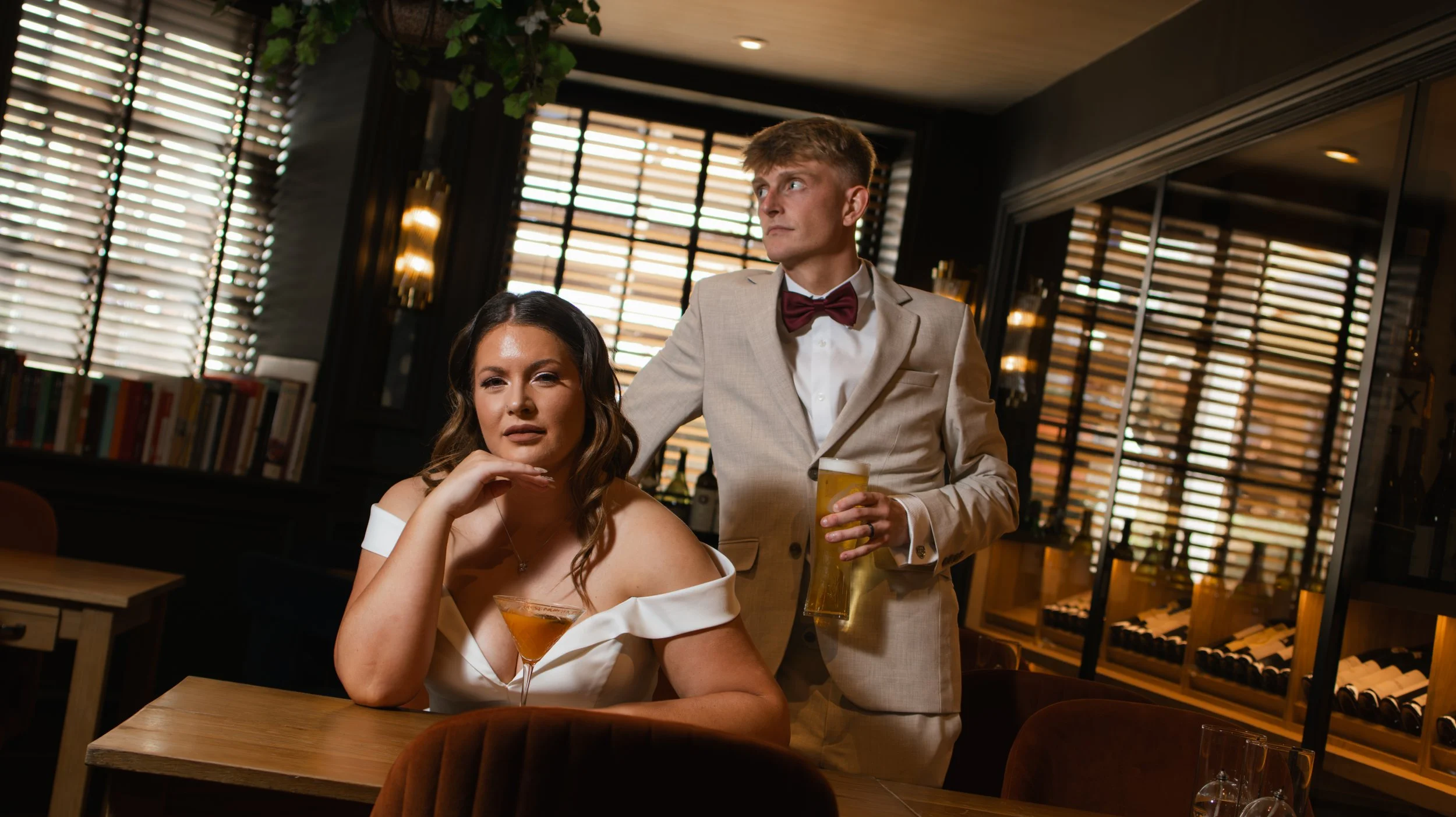 A woman sitting at a restaurant table with a drink in a martini glass and a man standing beside her holding a beer. The woman appears contemplative, resting her chin on her hand, while the man looks off to the side. The background features wooden blinds and bookshelves, creating a cozy, upscale atmosphere.