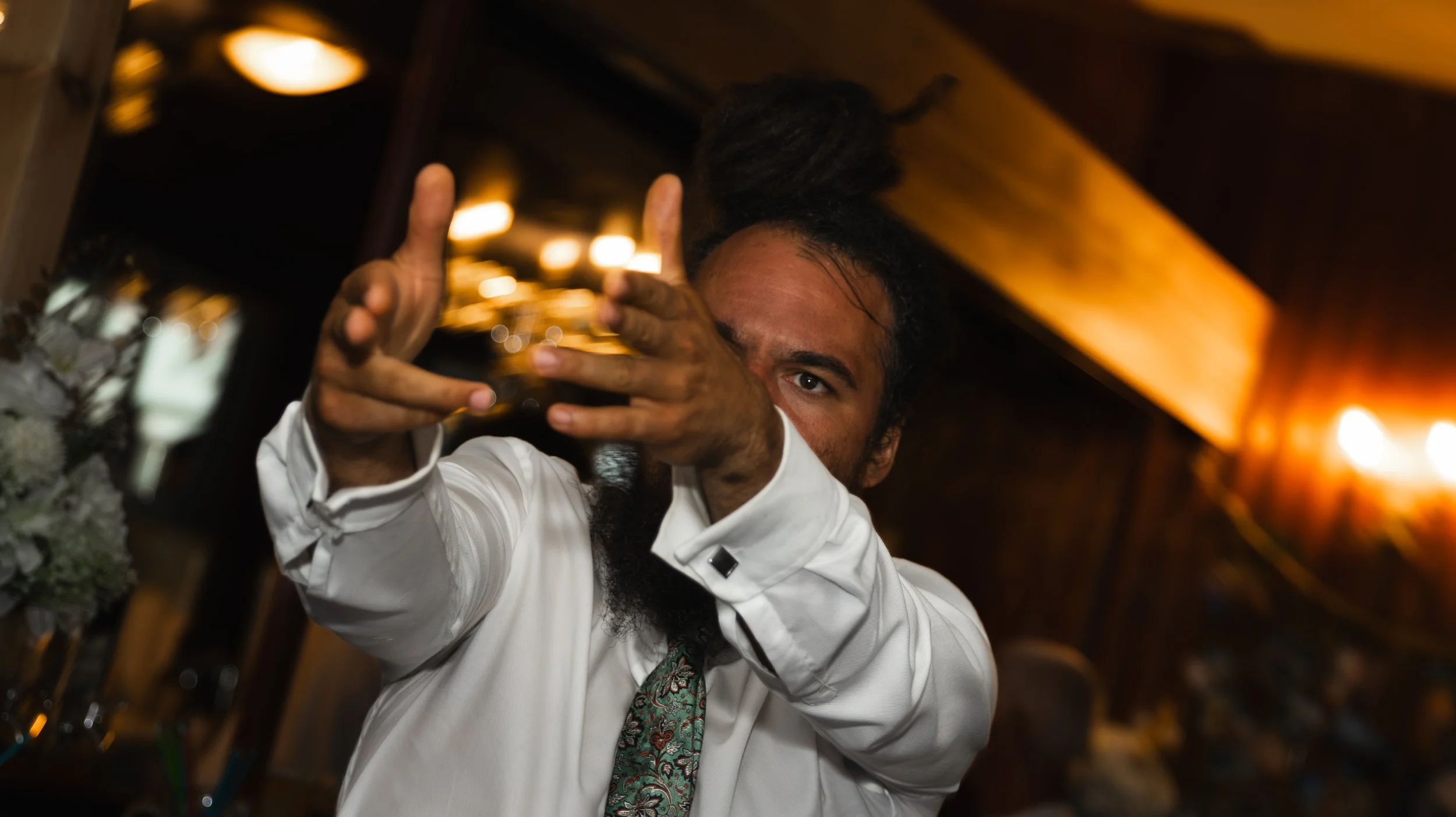 A man with a dark beard and moustache wearing a white shirt and a patterned tie, is gesturing with his hands and taking a photograph or video with a smartphone in a dimly lit, Warm, elegant setting.