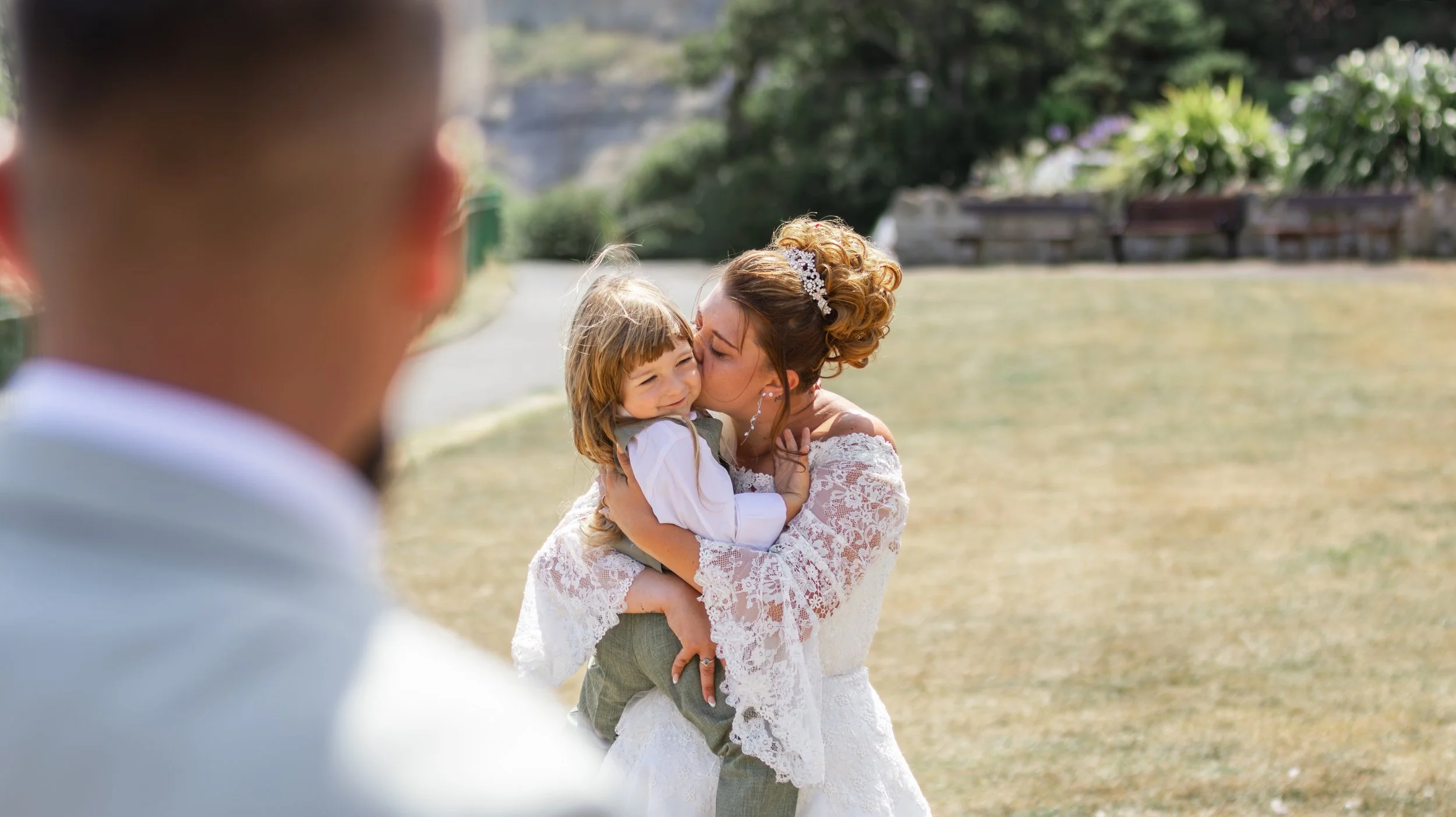 A bride in a lace wedding dress hugs a young girl with red hair, who is smiling and crying tears of happiness, during an outdoor wedding ceremony.