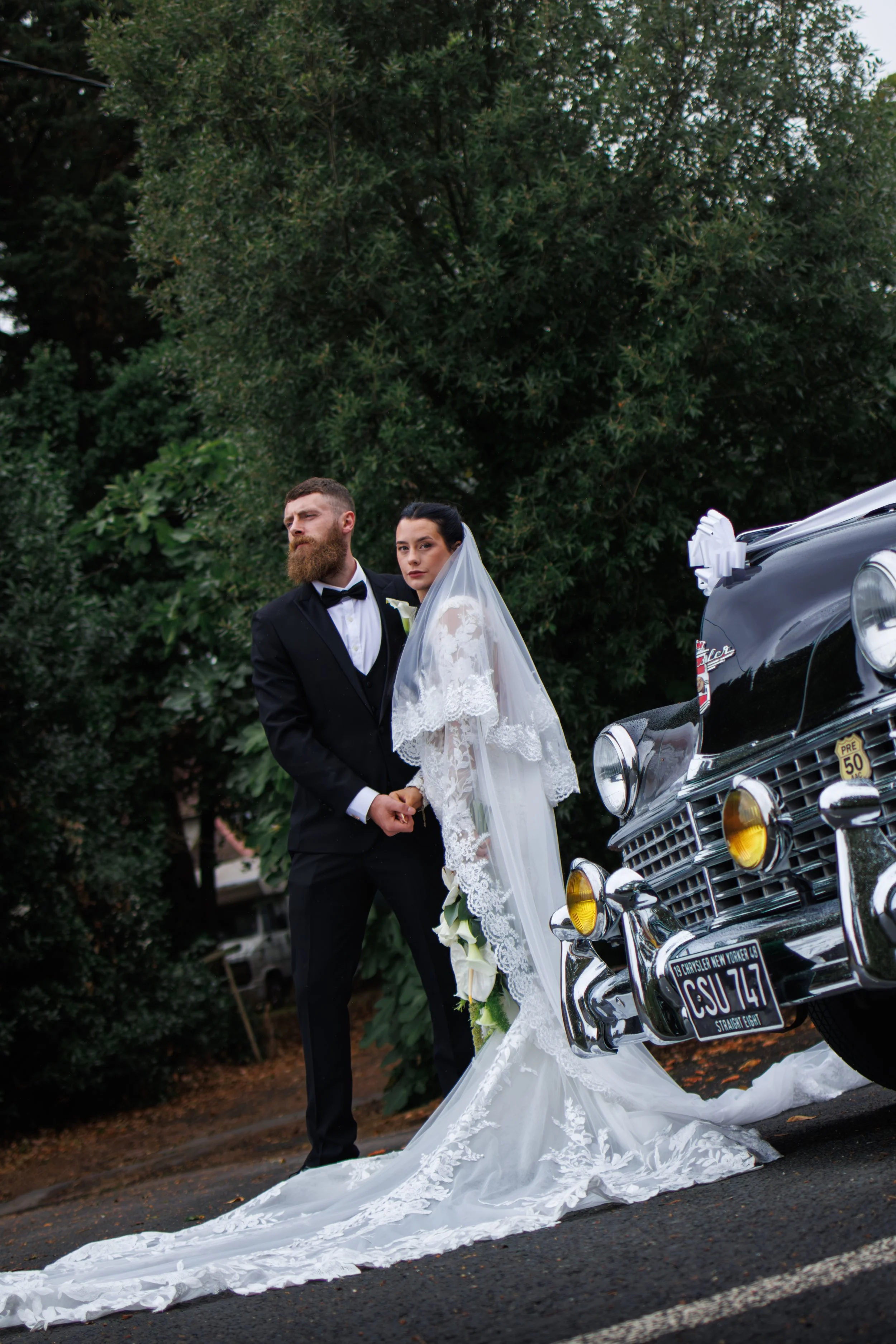 A bride and groom standing near a vintage black Cadillac decorated for a wedding, with lush green trees in the background.
