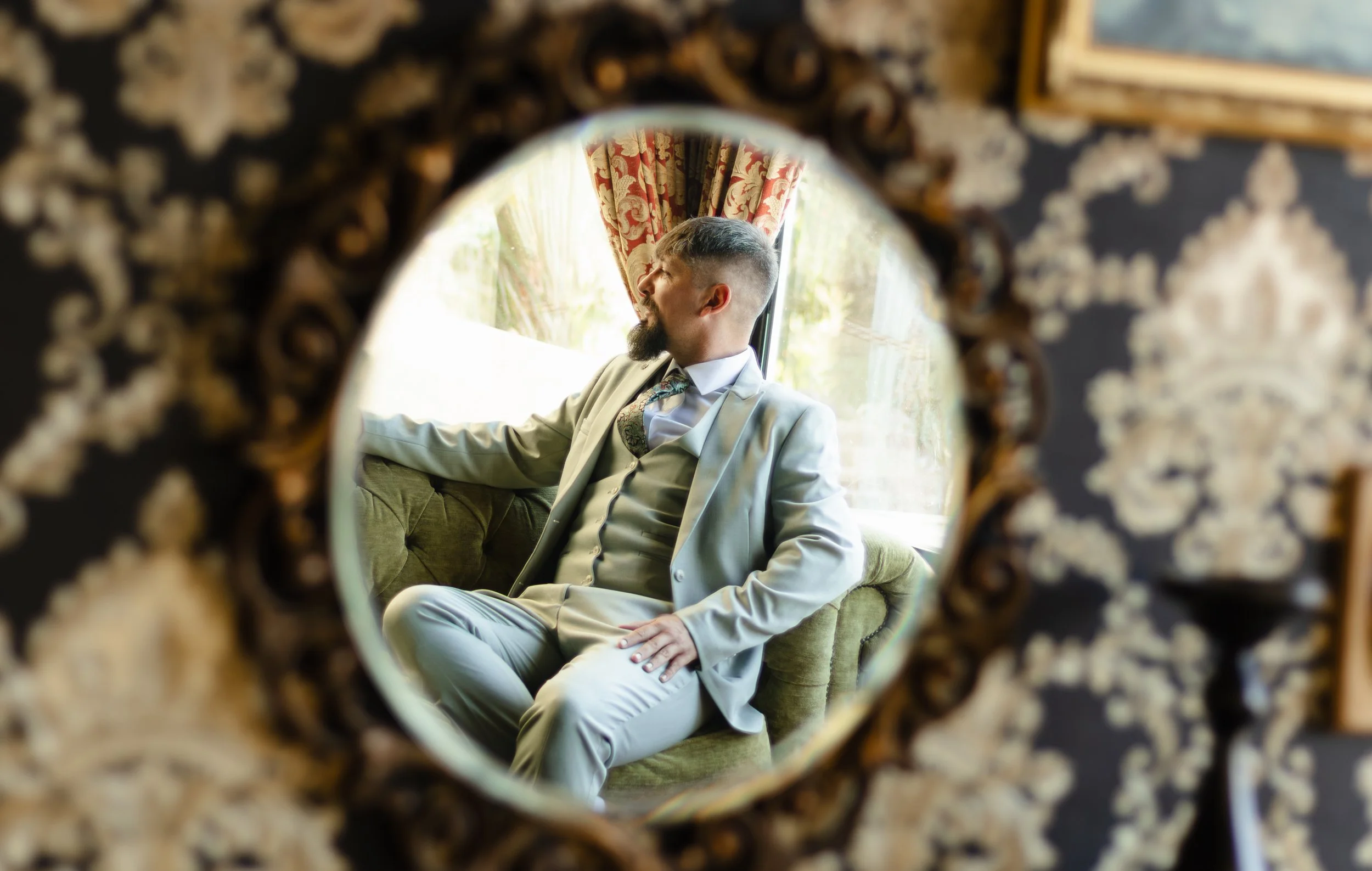 Man with a beard and mustache, wearing a light grey suit and floral tie, sitting on a green velvet sofa, seen through a decorative frame with ornate patterns, in front of a window with red floral curtains.
