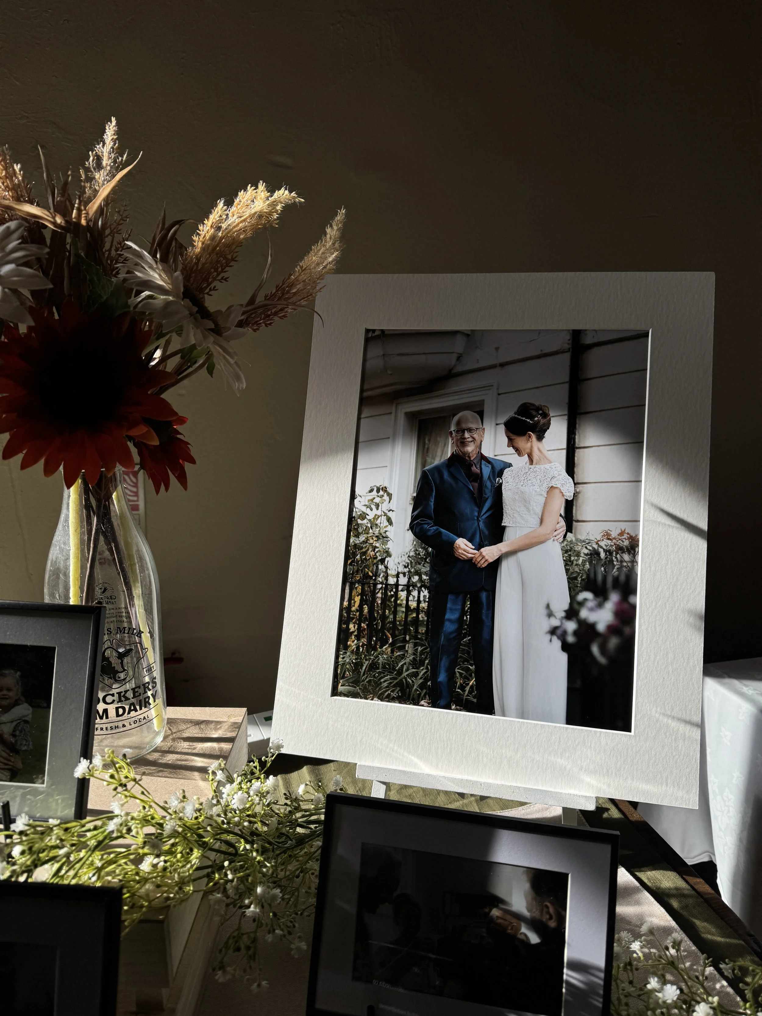A framed wedding photograph of a man and a woman smiling and holding hands, standing outside a house with white siding, surrounded by plants and flowers.