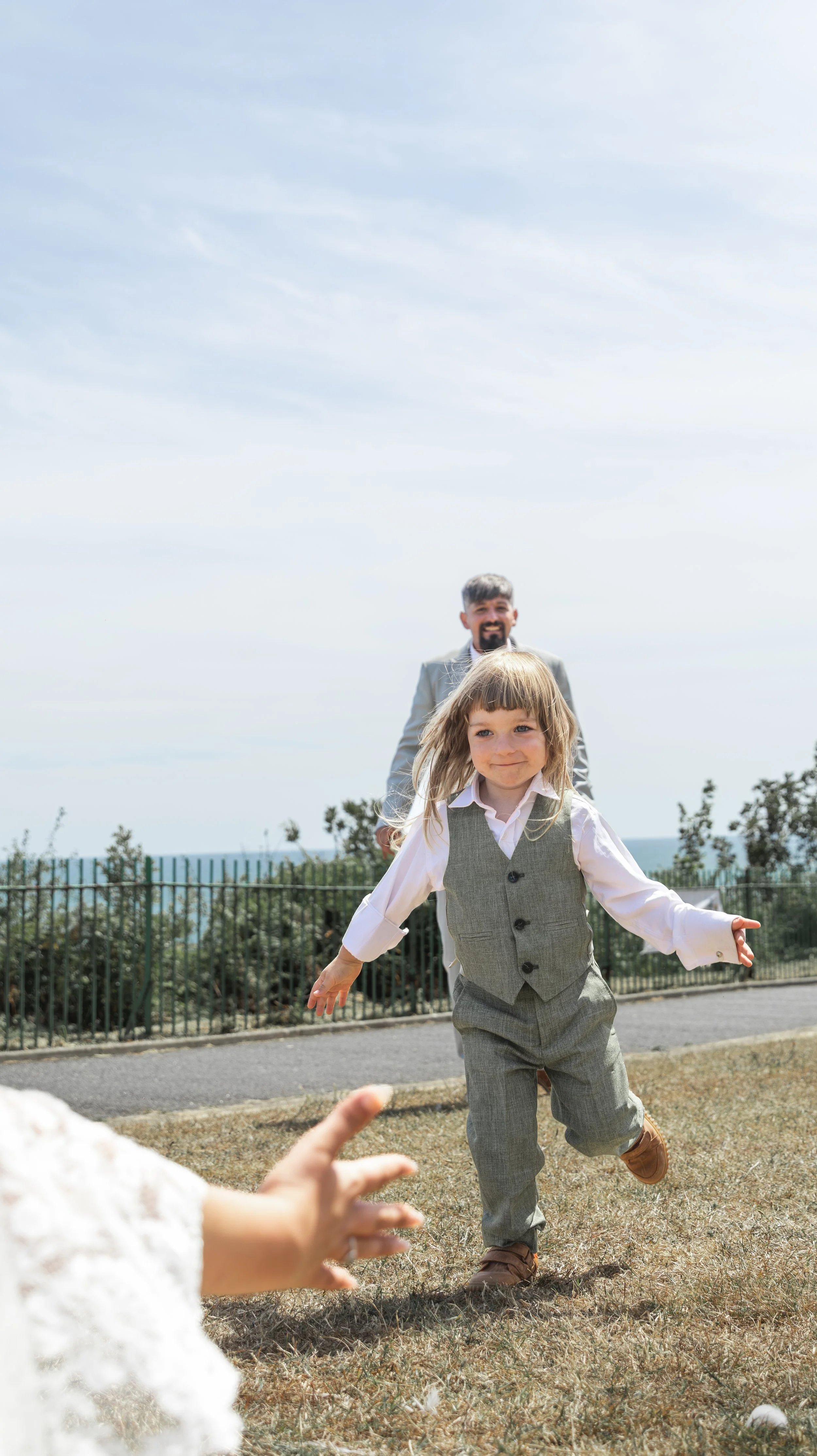 A young girl running outdoors towards a woman with open arms, with a man smiling in the background, on a sunny day near a fence and trees.