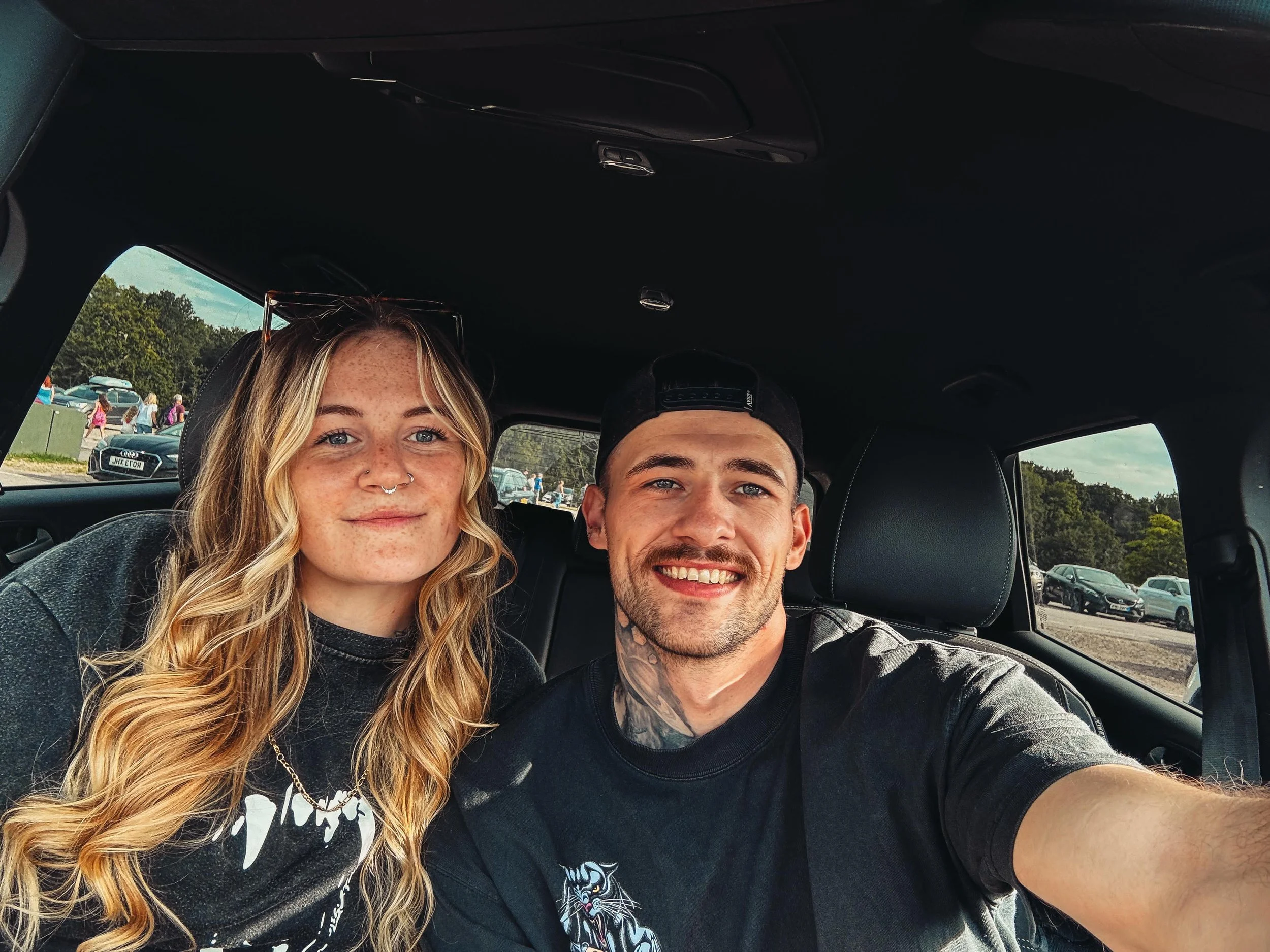 Two young adults, a blonde woman with freckles and a nose piercing, and a man with a beard and tattoos, smiling while sitting inside a car. Outside the car window, there are parked cars and a parking lot with people and trees in the background.