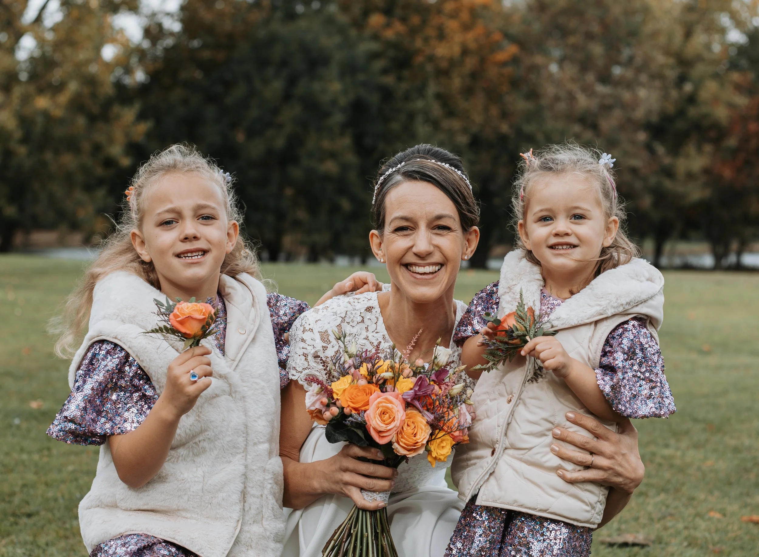 Four women, two young girls and one adult woman, enjoying an outdoor gathering. The women are smiling and holding bouquets of flowers, with trees and a grassy park in the background.