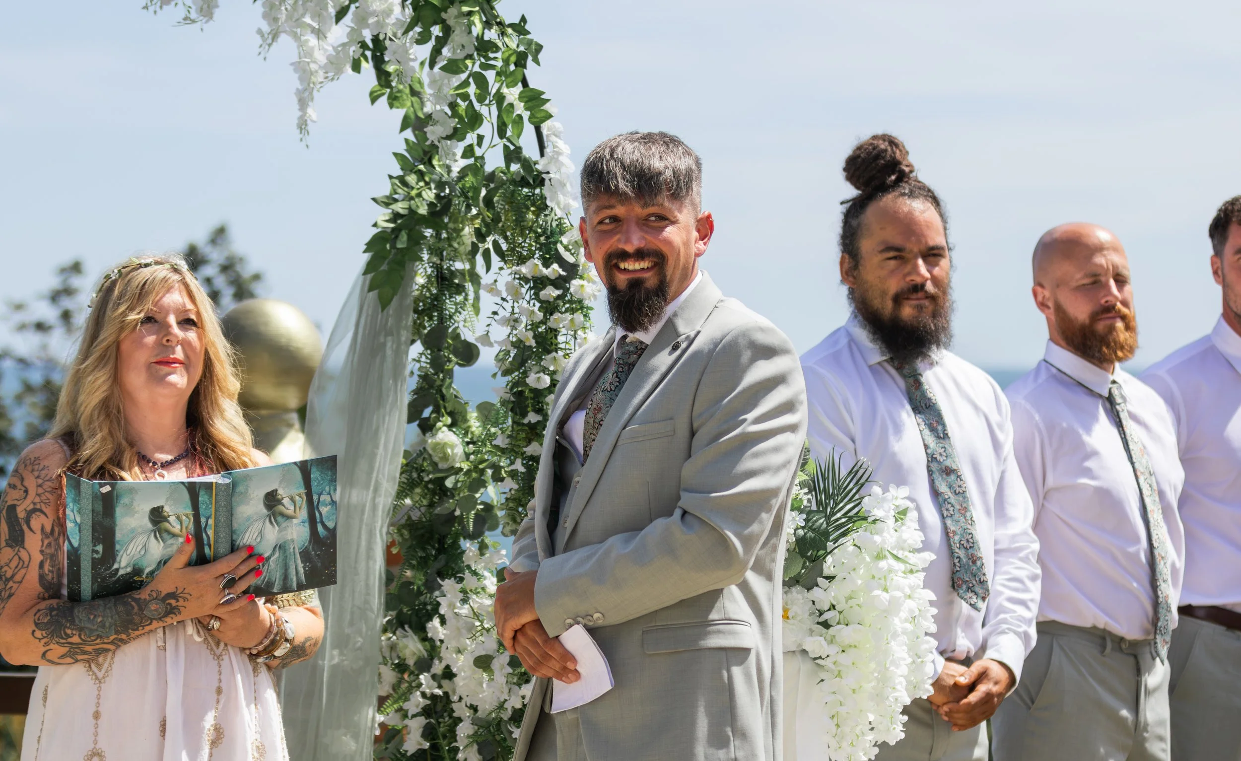 A wedding ceremony outdoors with four people standing in front of a floral arch. A woman with long blonde hair and tattoos on her arms is reading from a book, while three men stand to her right, wearing white shirts and patterned ties. They are all f