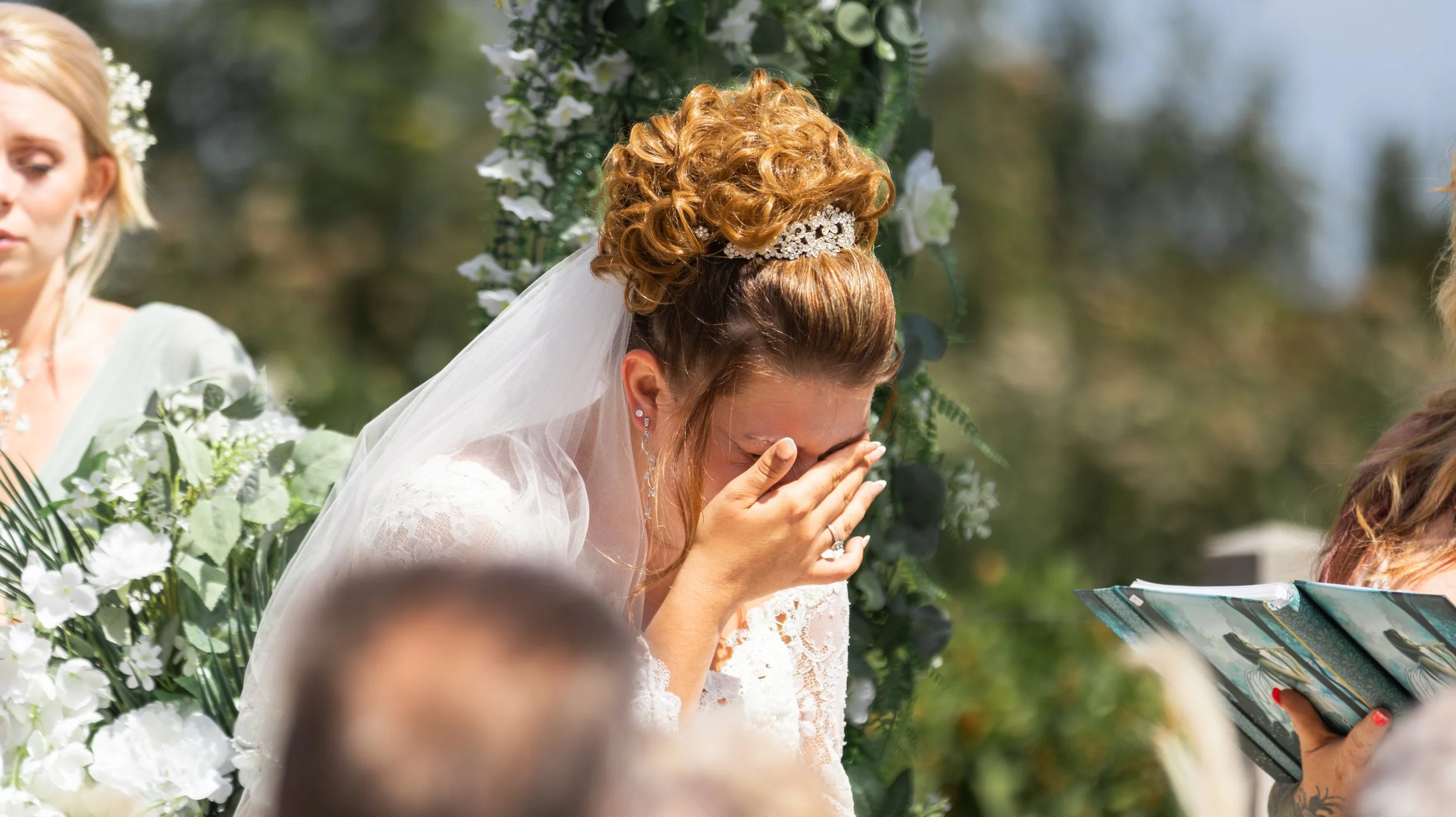 A bride with red hair, wearing a tiara and a white veil, covering her face with her hands, during outdoor wedding ceremony. She is dressed in lace, and floral decorations are visible in the background.