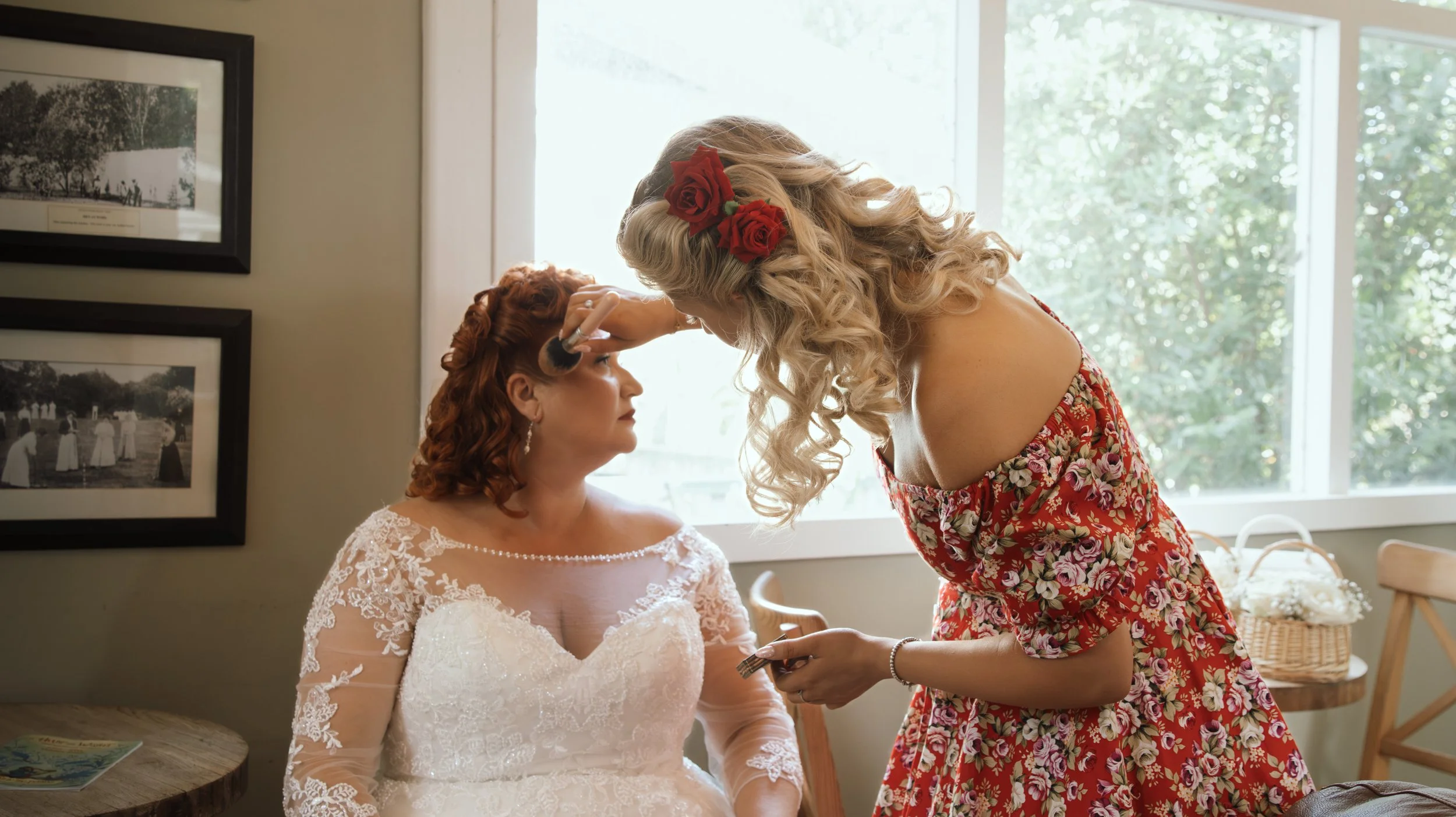 A bride with red hair and a lace wedding dress is having her makeup done by a woman with long curly blonde hair wearing a floral dress with red roses in her hair, in a room with large windows and framed black-and-white photographs on the wall.