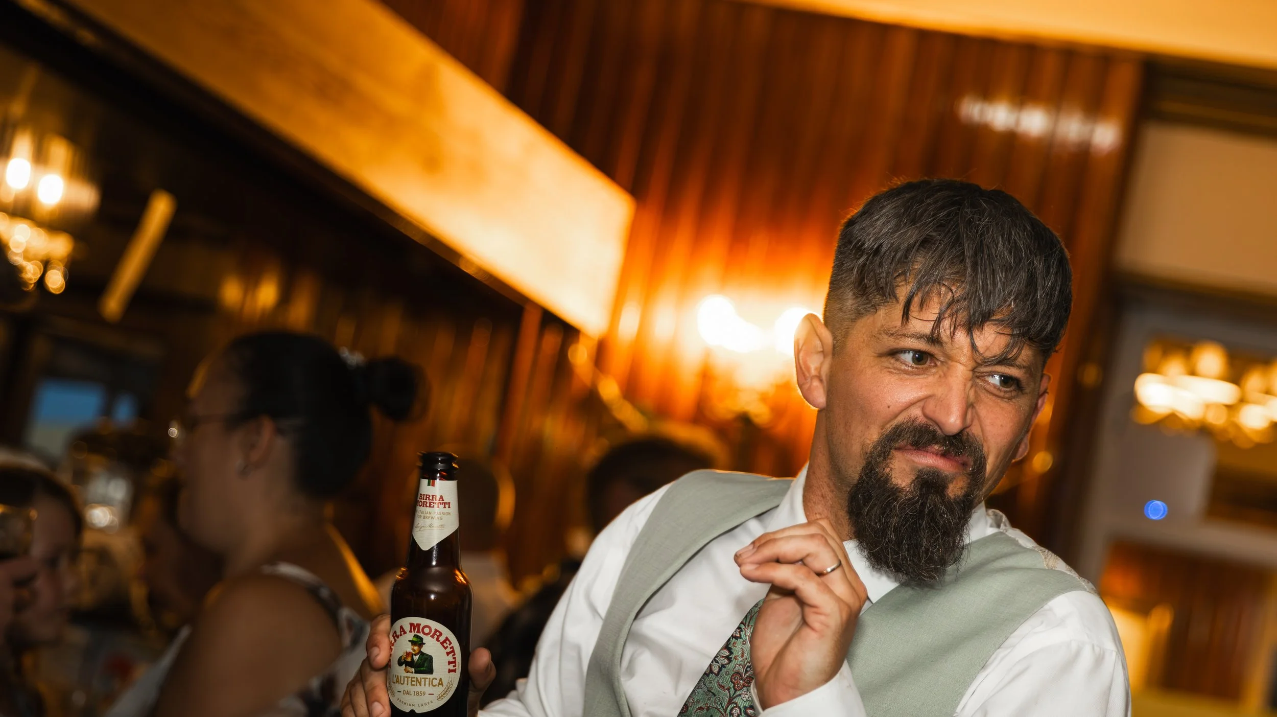 A man with dark hair and a beard, wearing a white shirt and vest, at a bar or restaurant with wooden walls and warm lighting, holding a beer bottle.